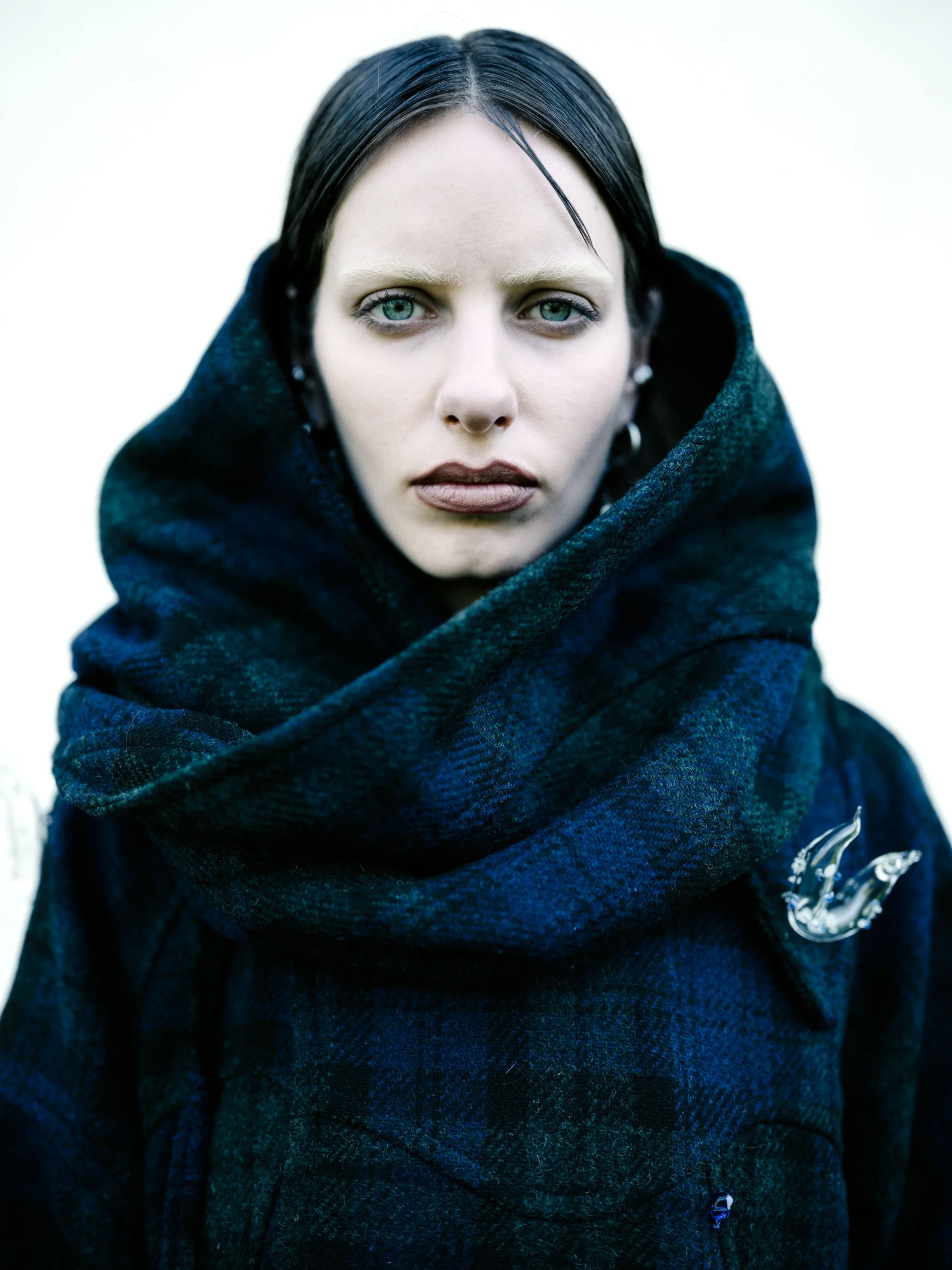 Nahaufnahme eines jungen Mannes mit blauen Augen und schwarzen Haare, trägt einen dunklen, karierten Schal und eine dunkle Jacke, im Hintergrund unscharf viel Weiß