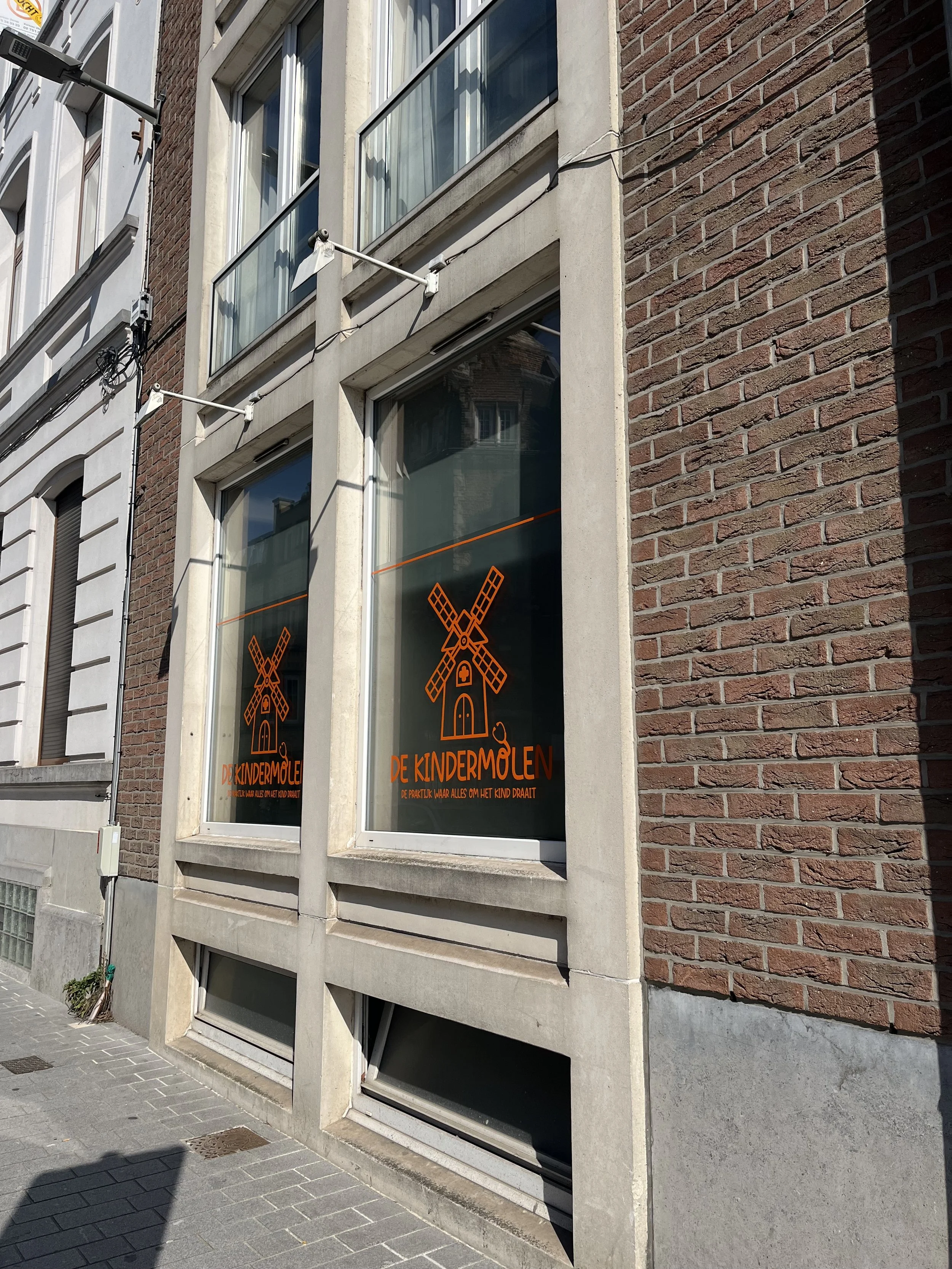 Gezicht op een gebouw met drie grote ramen die reclame voor 'De Kindermolen' tonen, een kleurrijk kinderbedrijf met een molen als logo, op een straat met een trottoir.