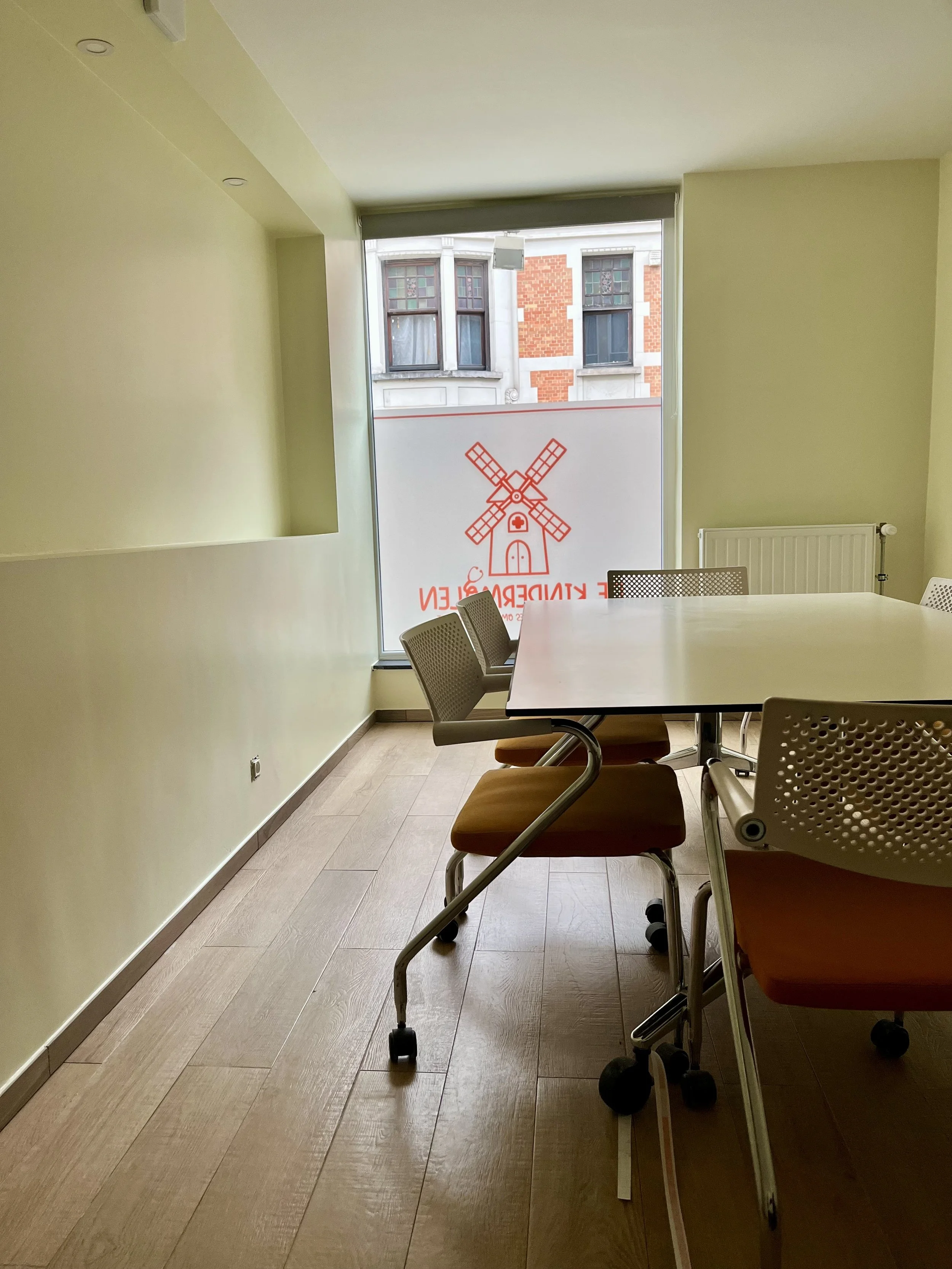 Vergadertafel met vier stoelen in een lichtgroene kamer met groot raam en gevel van een ouder gebouw buiten.