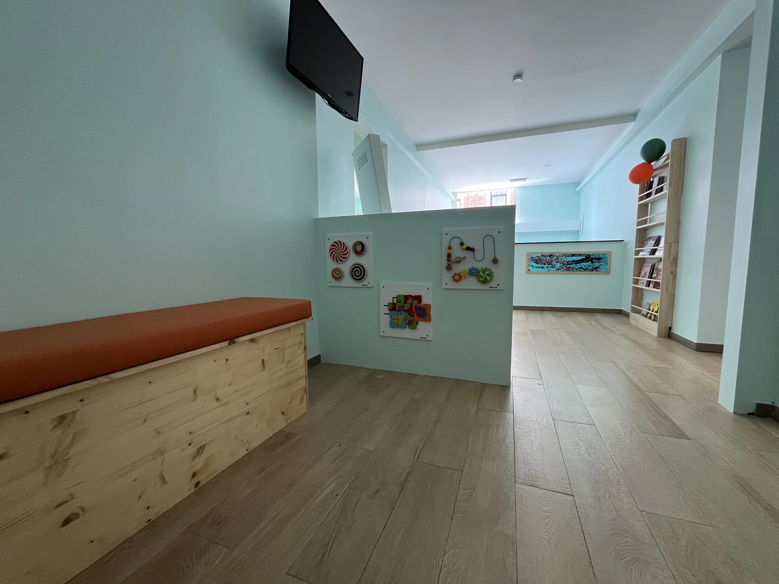 Binnenruimte met houten vloer, lichte mintgroene muren, een plankenbank met oranje zitkussen, een zwarte televisie aan de muur, en een scheidingswand met speelgoed en decoraties. In de achtergrond een boekenrek met ballonnen. Gedeeltelijk zichtbaar daglicht door een raam.