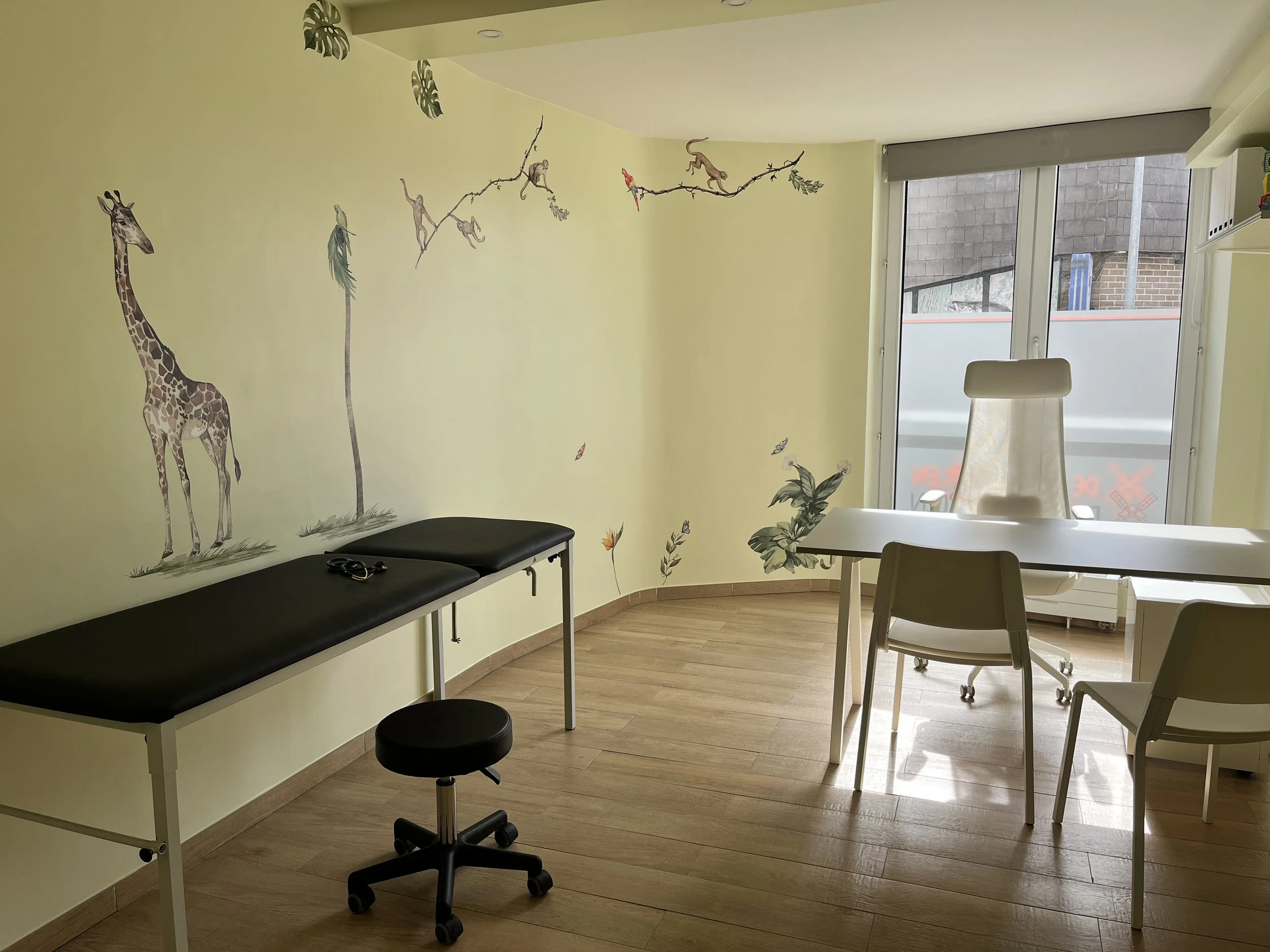 Woonkamer of kantoorruimte met gele muur met jungle muurschildering, witte tafel en bureaustoel voor ramen met uitzicht op een gebouw met bakstenen