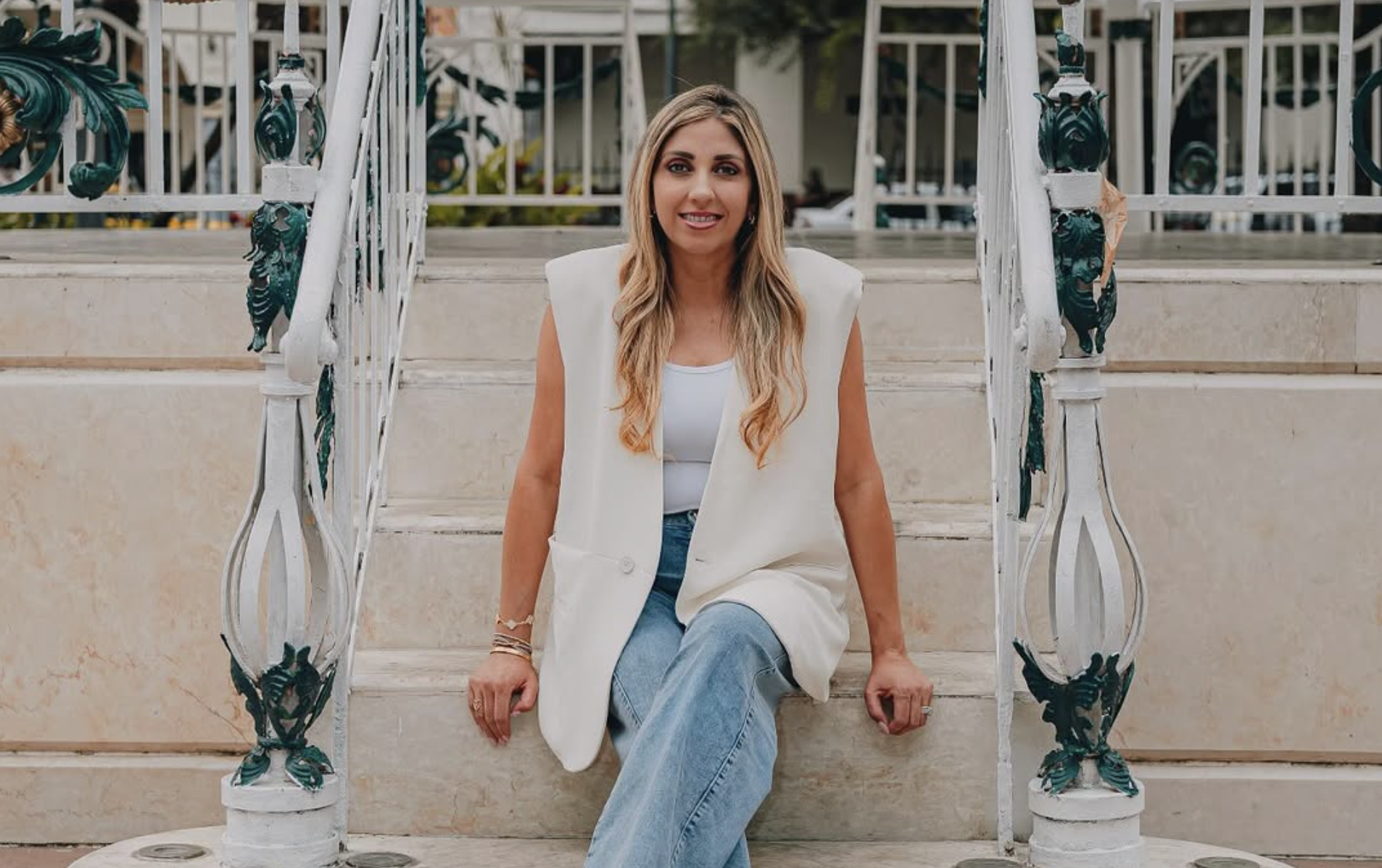 A woman with long blonde hair sitting on outdoor stairs, wearing a white sleeveless blazer and light blue jeans, smiling at the camera.