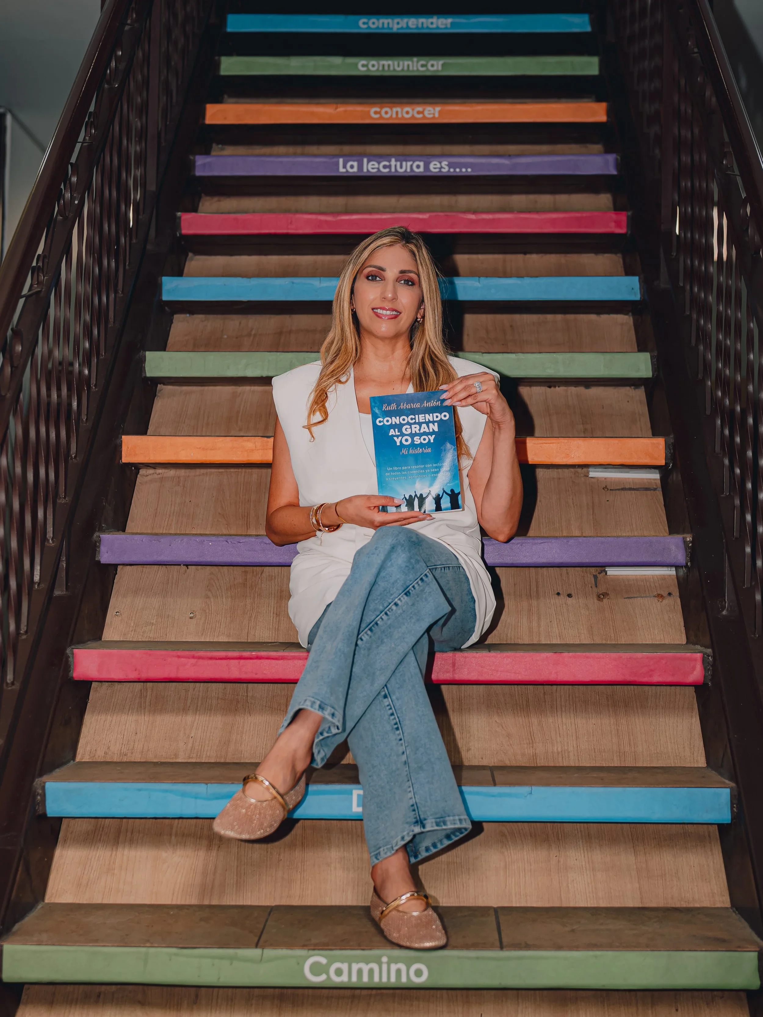 A woman sitting on colorful stairs holding a book titled "Conociendo al Gran Yo Soy" in Spanish, with the staircase labeled with Spanish words such as "compreder," "comunicar," "conocer," "La lectura es," and "Camino."