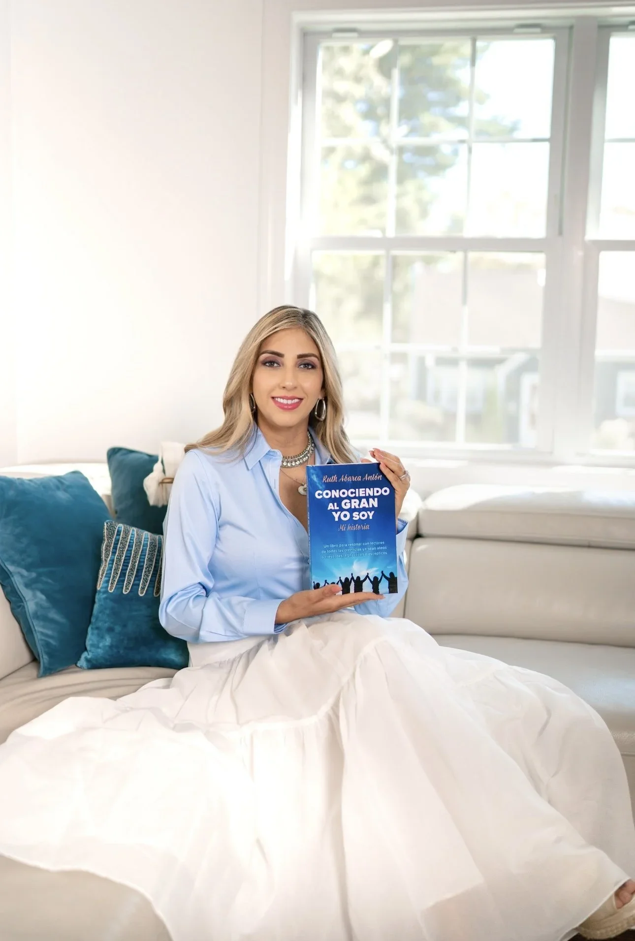 A woman with blonde hair, wearing a light blue blouse and a white skirt, sitting on a cream-colored sofa with blue pillows. She is holding a blue book titled 'Conociendo al Gran Yo Soy' and is smiling at the camera. Behind her is a large window with sunlight coming through.