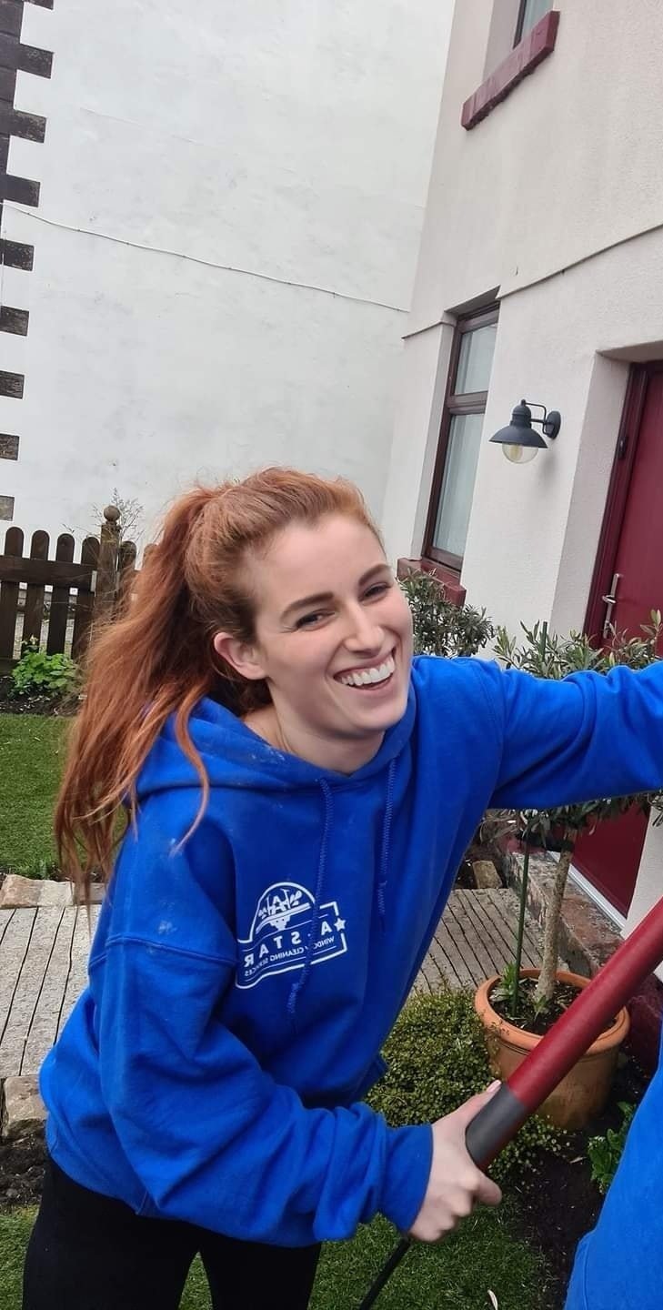 A young woman with red hair smiling while holding a garden tool in a backyard.