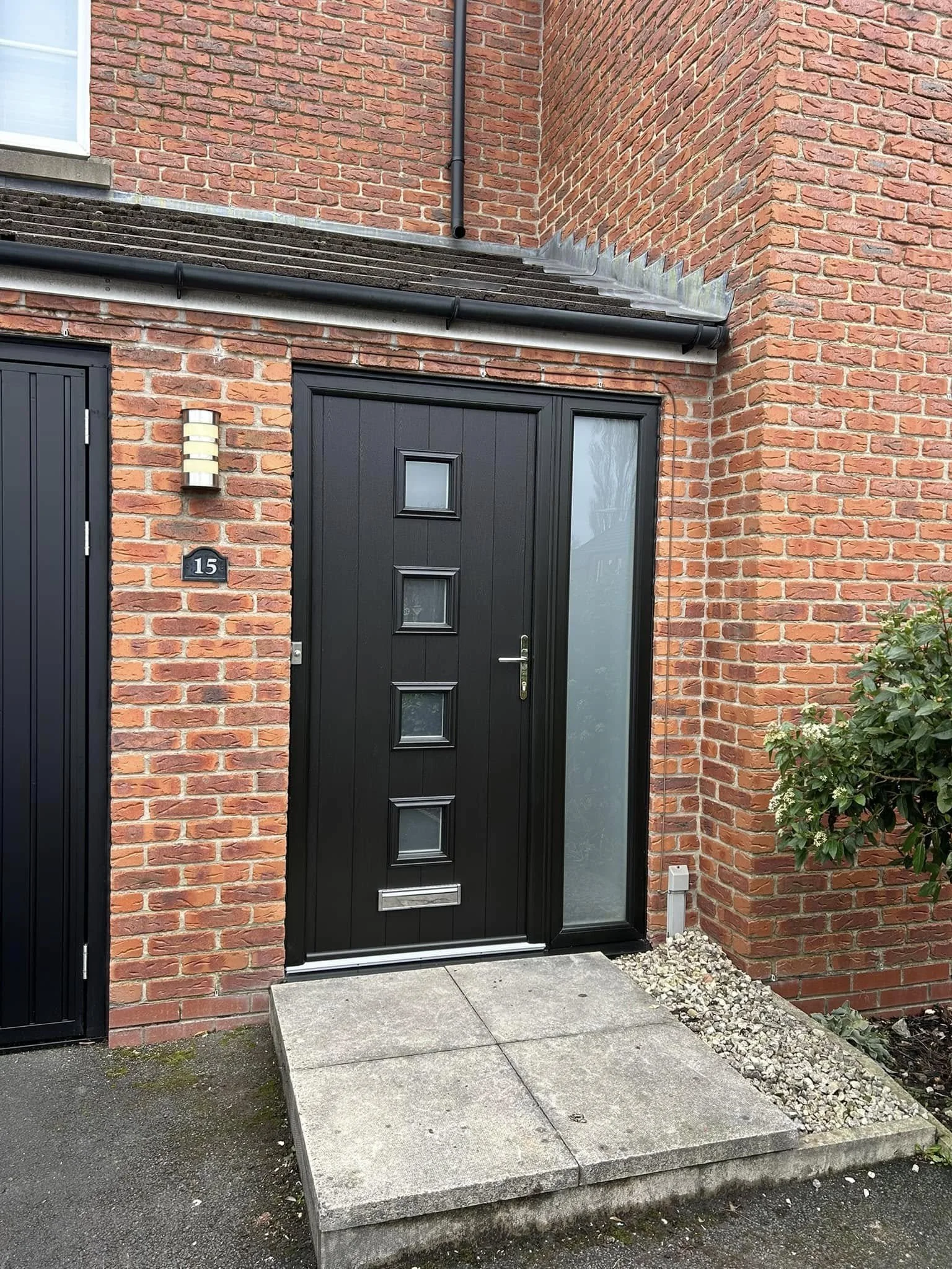 Black front door with small square windows on brick house exterior, with a concrete step leading to the entrance, and a small shrub on the right side.