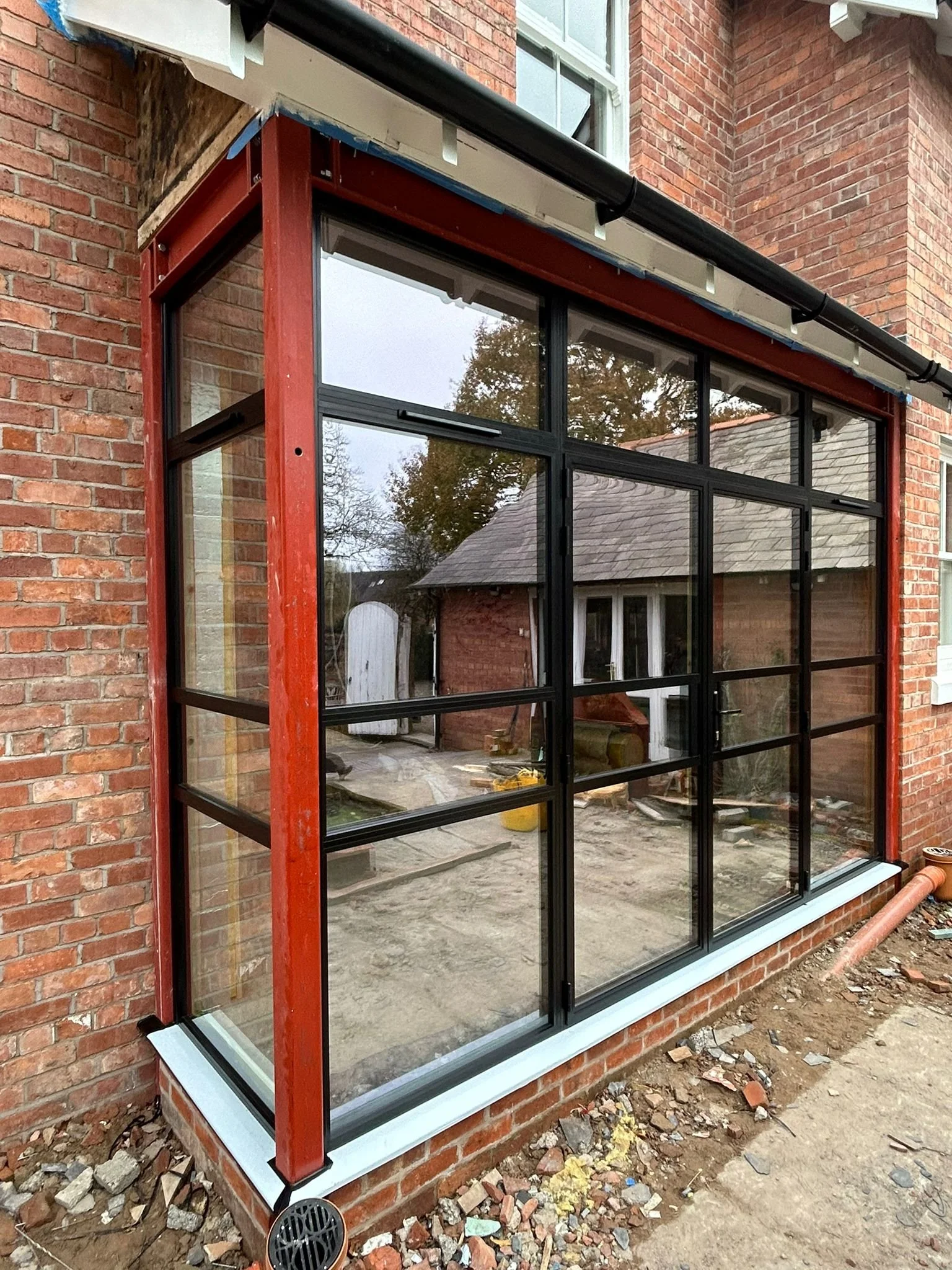 A newly installed enclosed porch with large glass windows framed in black, set against a brick house exterior, with construction materials and dirt on the ground.