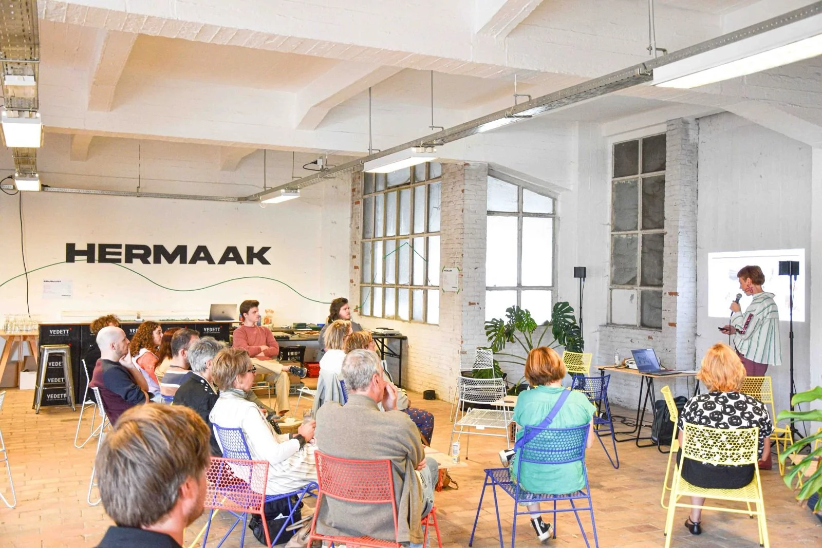 A woman giving a presentation to a seated audience in an industrial-style room with large windows, colorful chairs, and large plants.
