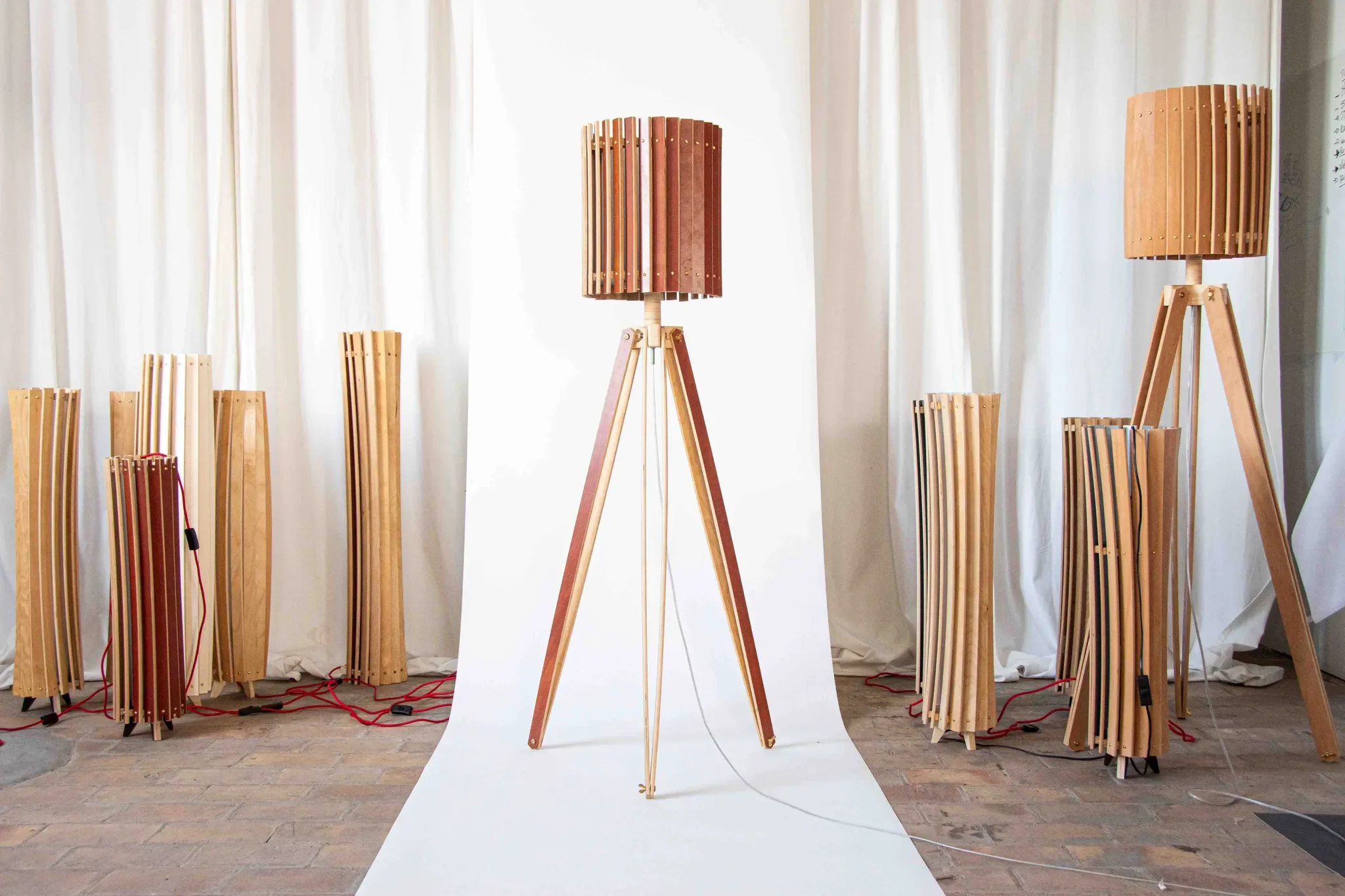 Display of various wooden lamps and lampshades on a brick floor with white curtains in the background.