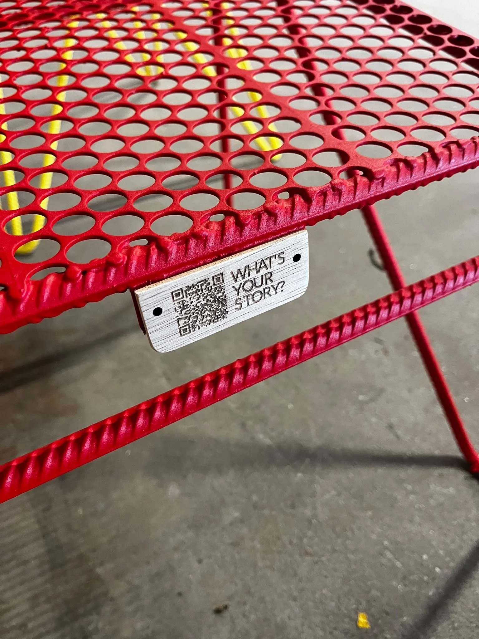 A red metal outdoor table, made from pre-concumer waste, with a perforated top surface and a wooden tag hanging from it. The table is on a concrete surface. The table has an object identity.