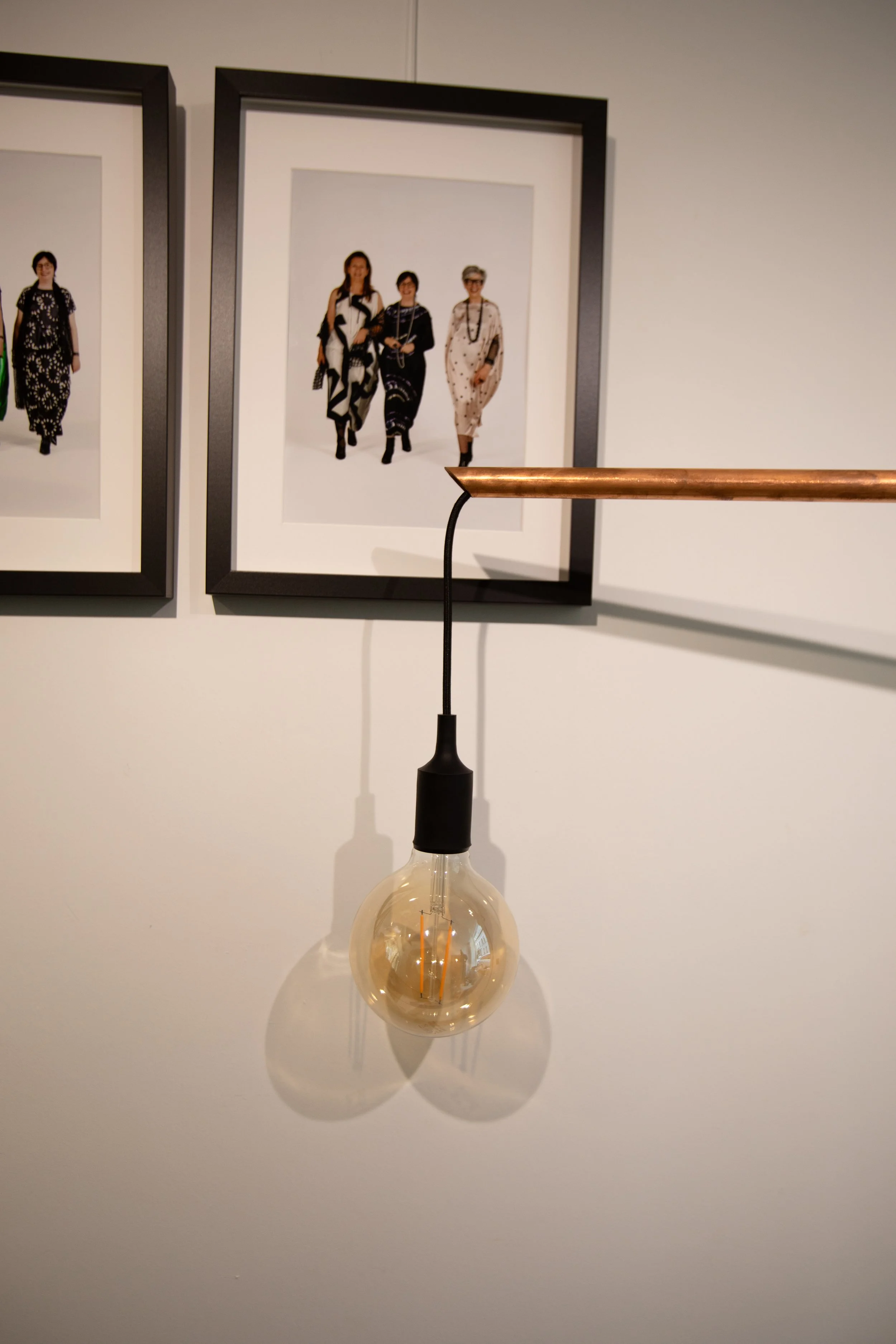 A hanging light bulb with a black socket, casting a shadow on a white wall with framed photographs of women in fashionable clothing.