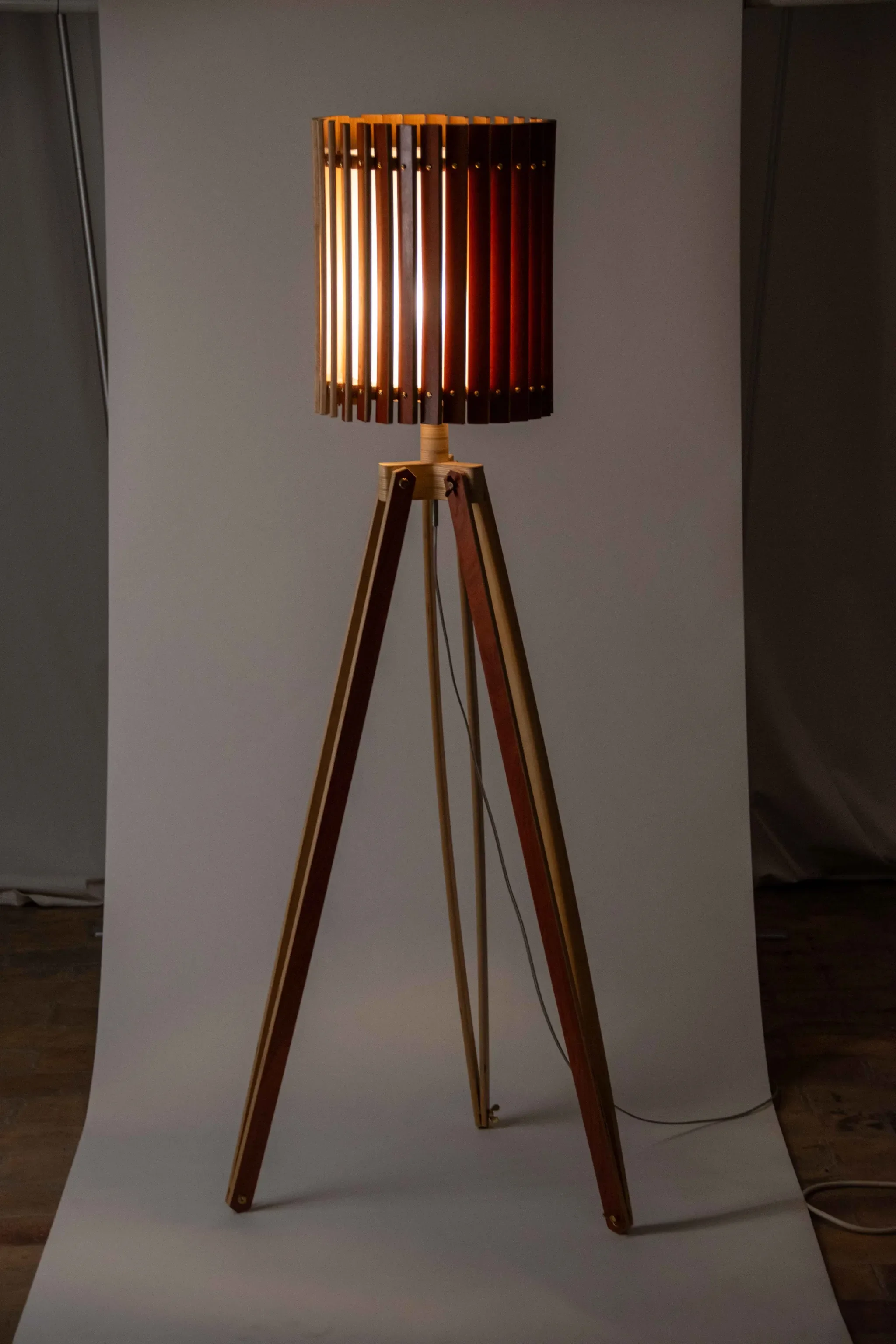 A standing floor lamp with a wooden tripod base and a cylindrical wooden slat lampshade emitting warm light.