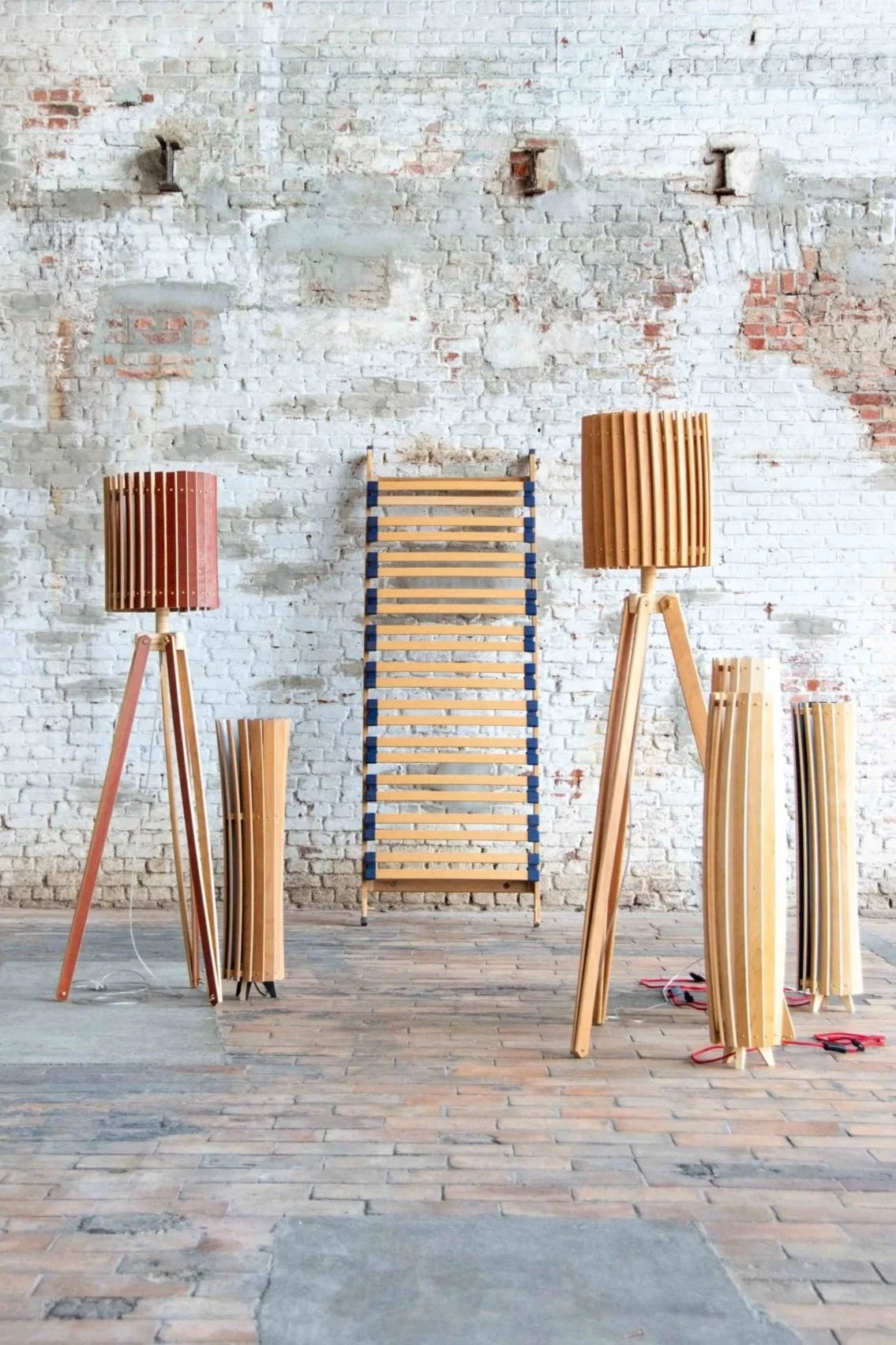 Multiple wooden lamps and stands arranged against a white brick wall in a room with a brick floor.