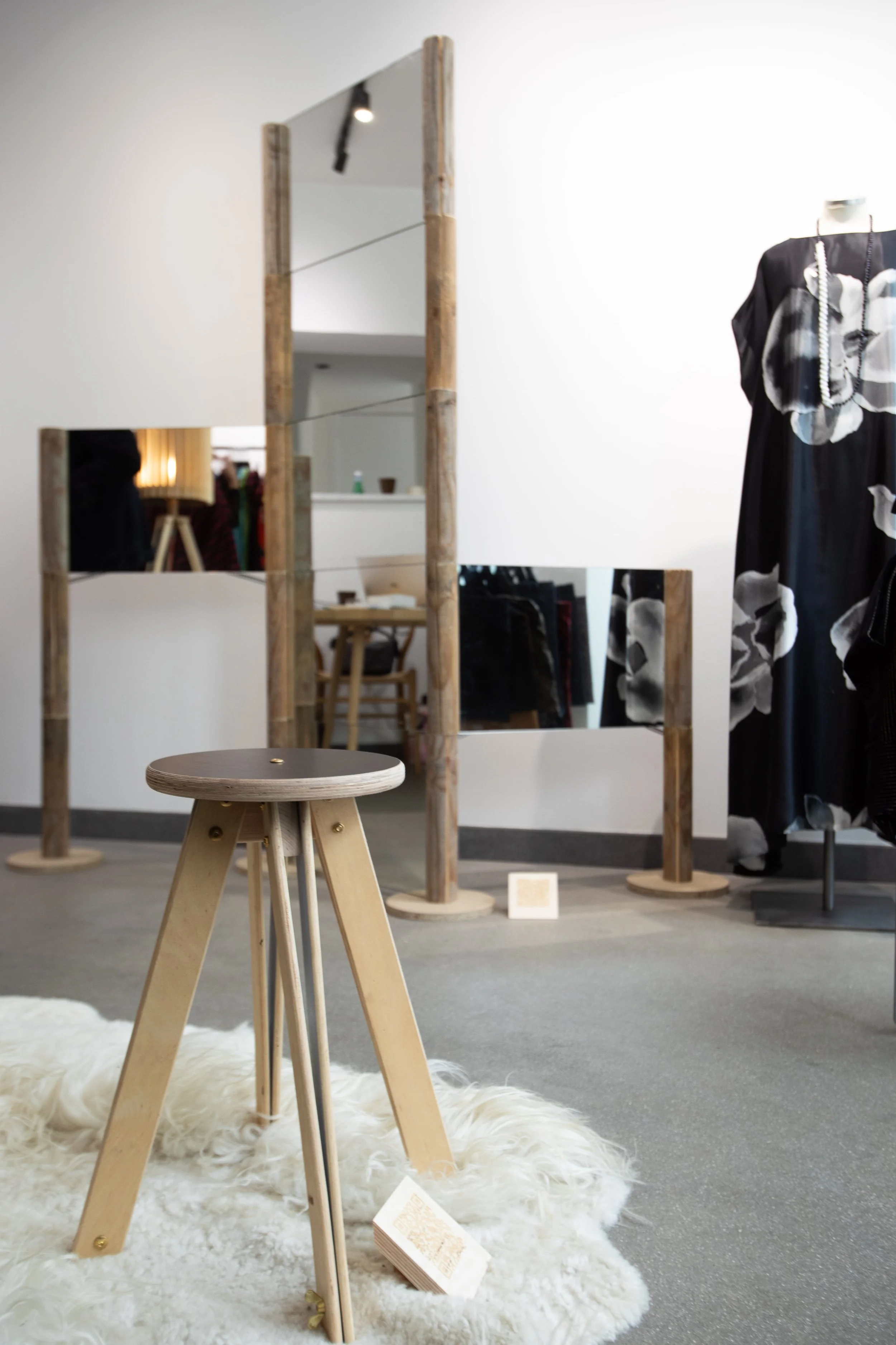 Interior of a retail store displaying furniture, mirrors, and clothing, with a rustic wooden and glass partition, a white fluffy rug, and a small wooden stool in the foreground.