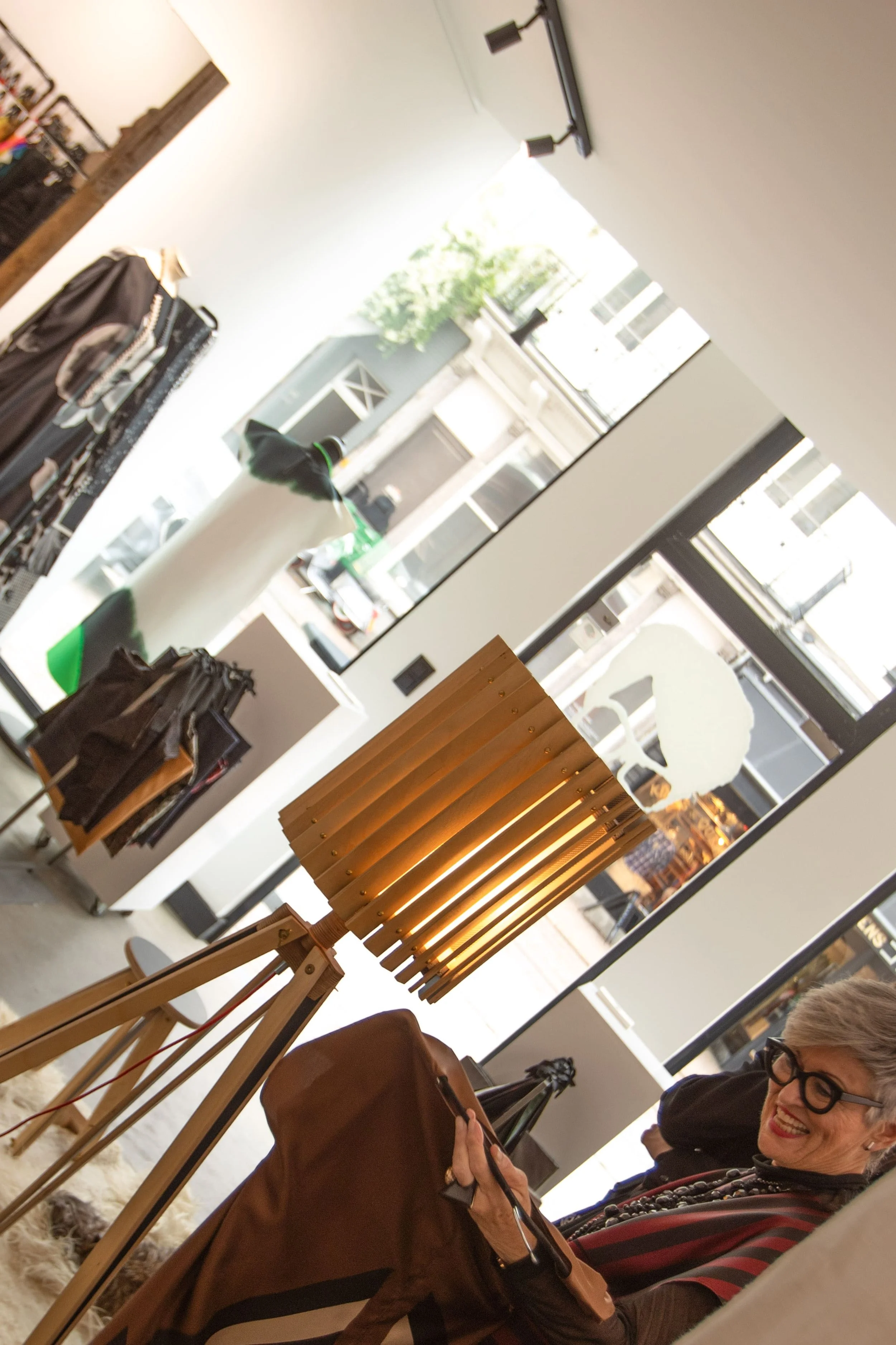 A woman with gray hair and glasses smiling inside a clothing store, sitting on a chair and holding a tablet. The store has large windows, a wooden lamp, and clothing displays.