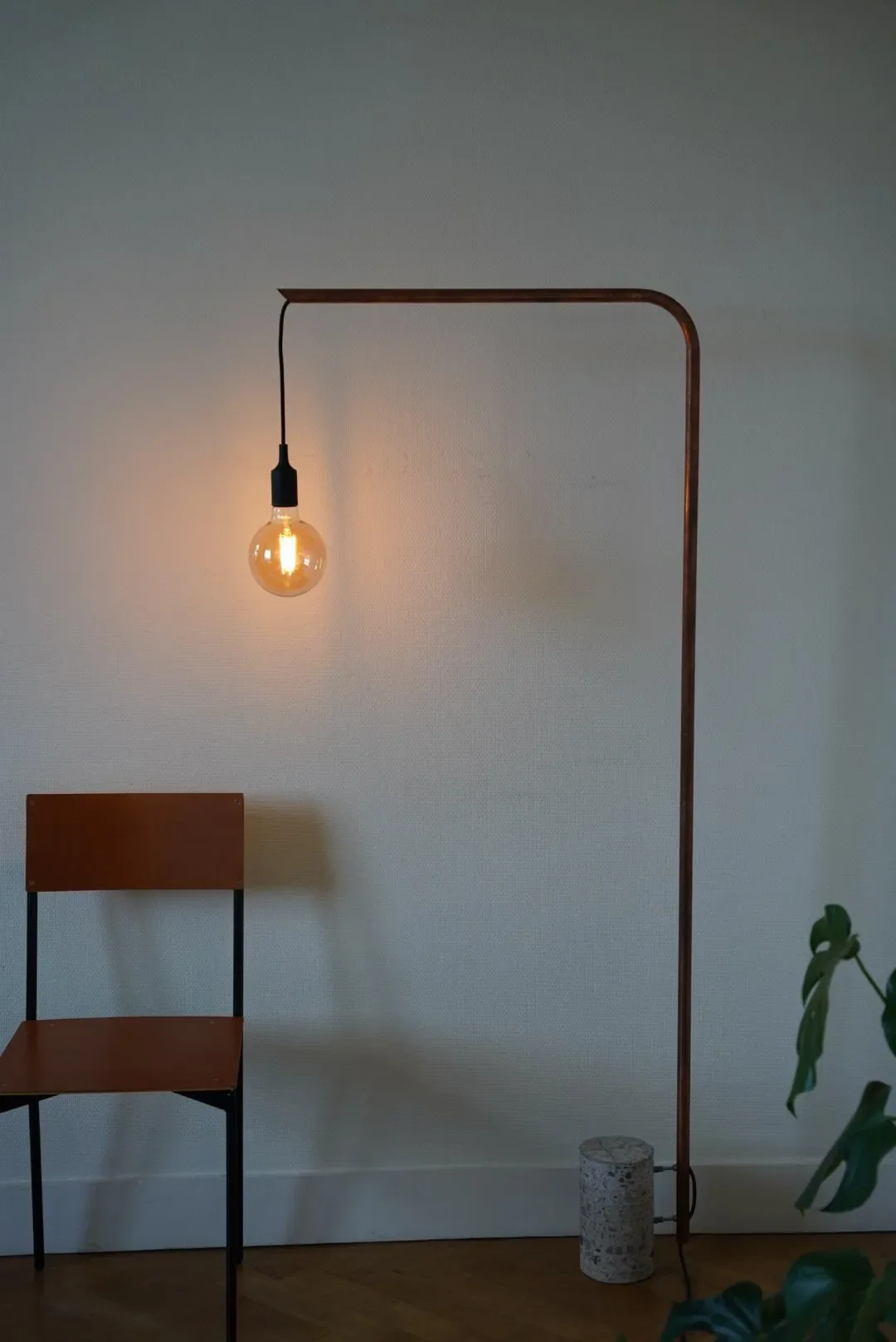 Interior room featuring a minimalist brown chair, a standing lamp with a black cord and small stone base, and a glowing Edison-style light bulb hanging from a copper-colored pipe structure, against a plain off-white wall with a small green plant partially visible on the right.