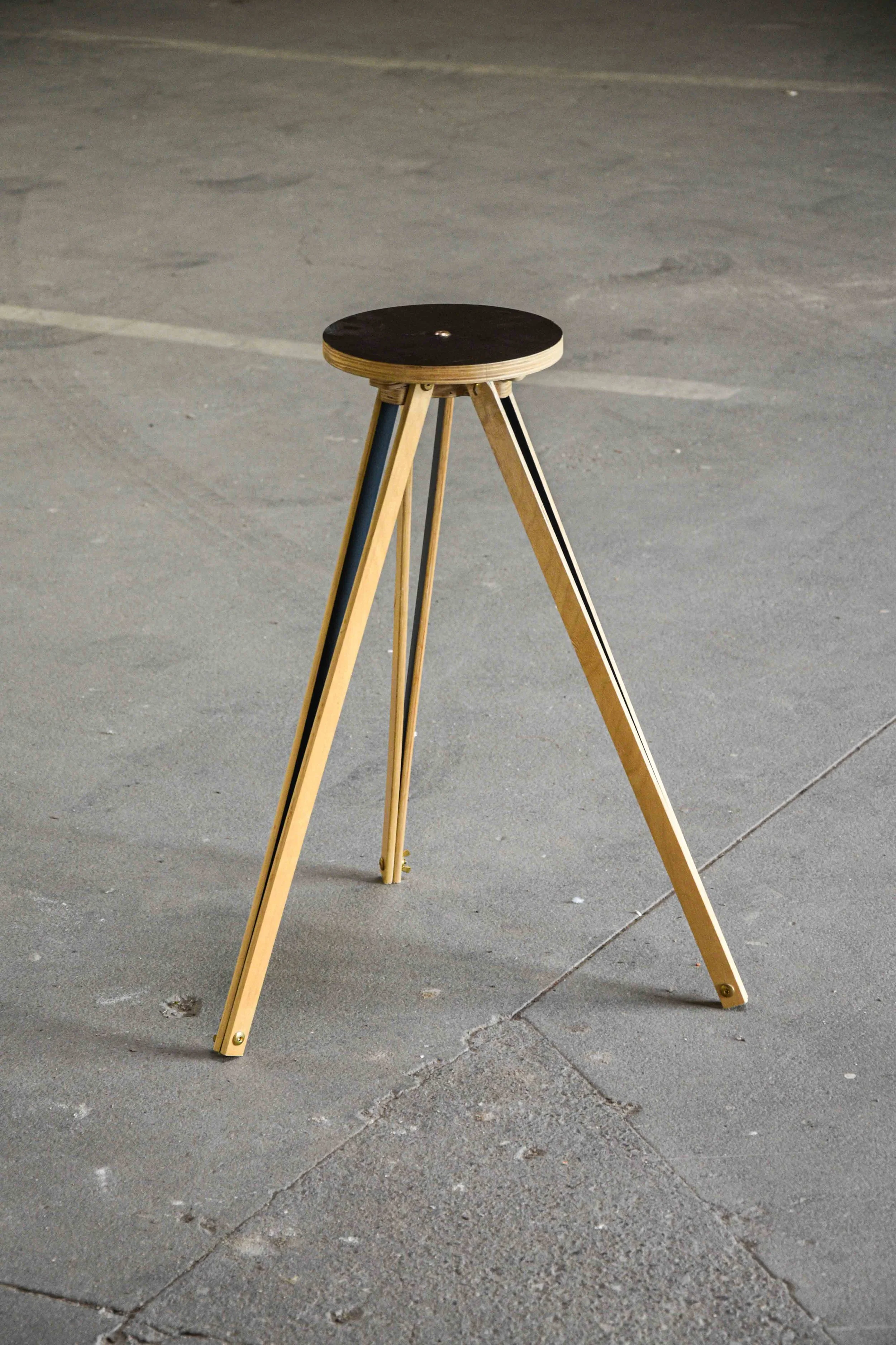 A wooden tripod stand with a round black top, placed on a concrete floor.