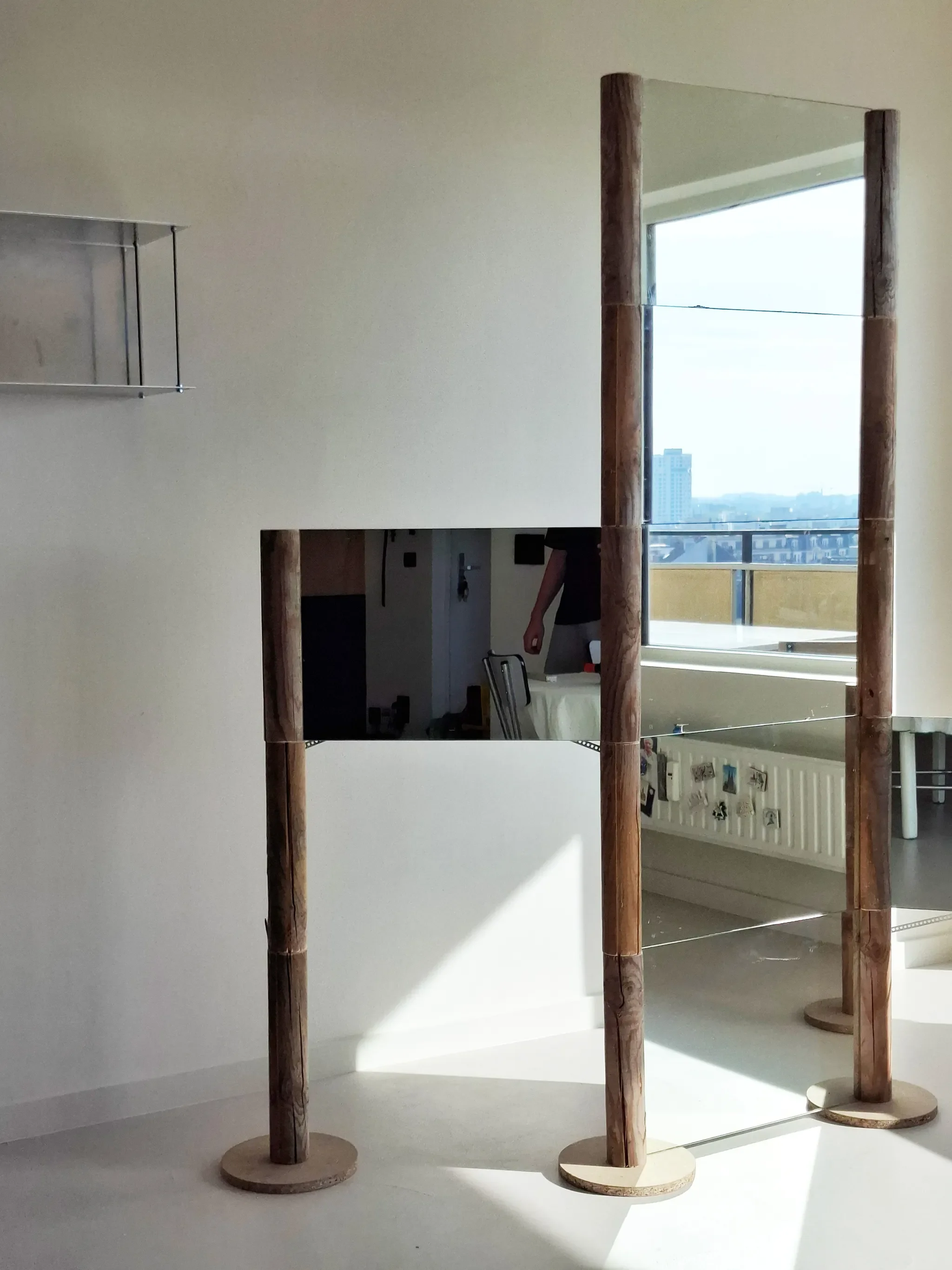 Interior view of a bright room with a large window showing a cityscape. A mirror with wooden pillars and horizontal wires in front is mounted on the wall, creating a reflective surface that reveals part of the room and a person’s arm.