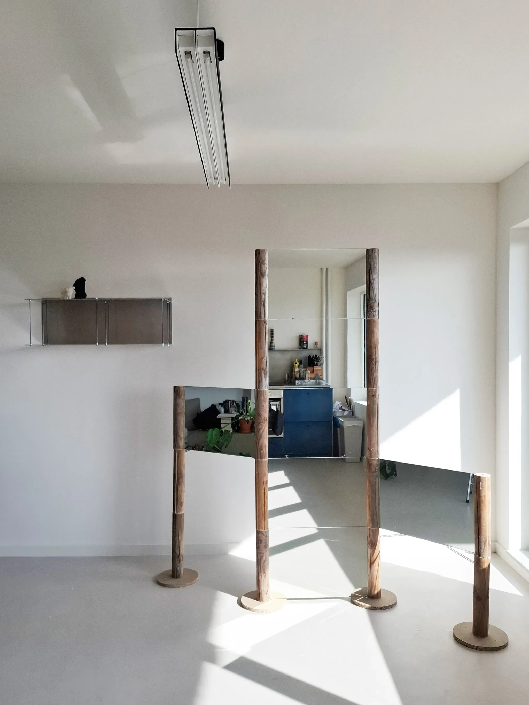 Minimalistic interior with a wooden rectangular mirror and a wooden-frame side panel, illuminated by natural sunlight, with a kitchen area in the background.