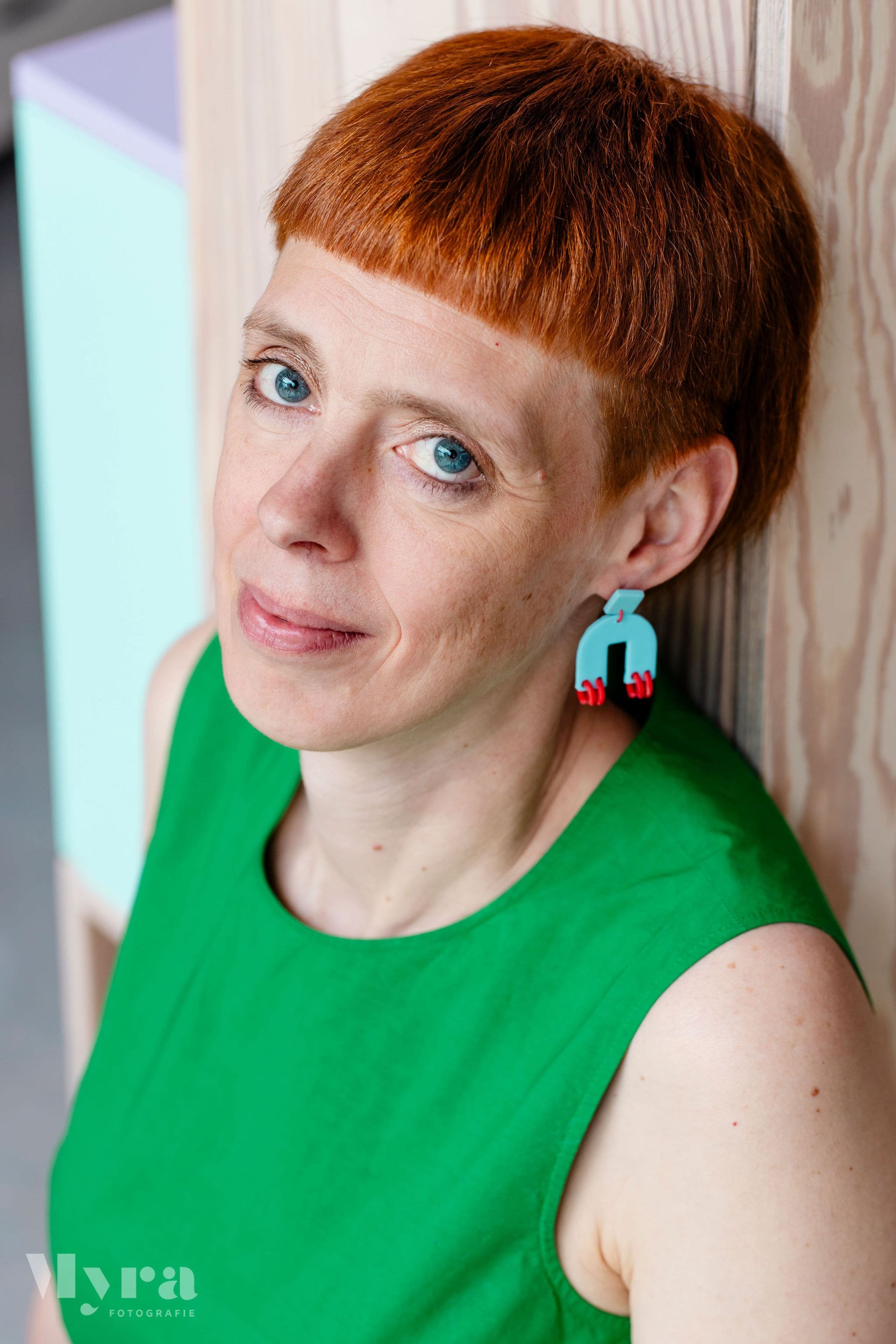 A woman with short red hair, blue eyes, wearing a sleeveless green top and blue earrings, leaning against a wooden wall.