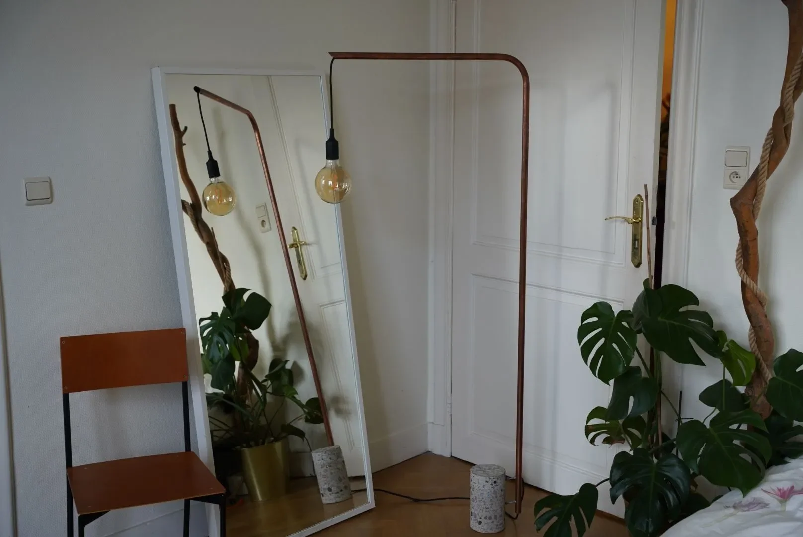 Room with a large standing mirror, a chair, two hanging lightbulbs, two potted plants, and a closed door.