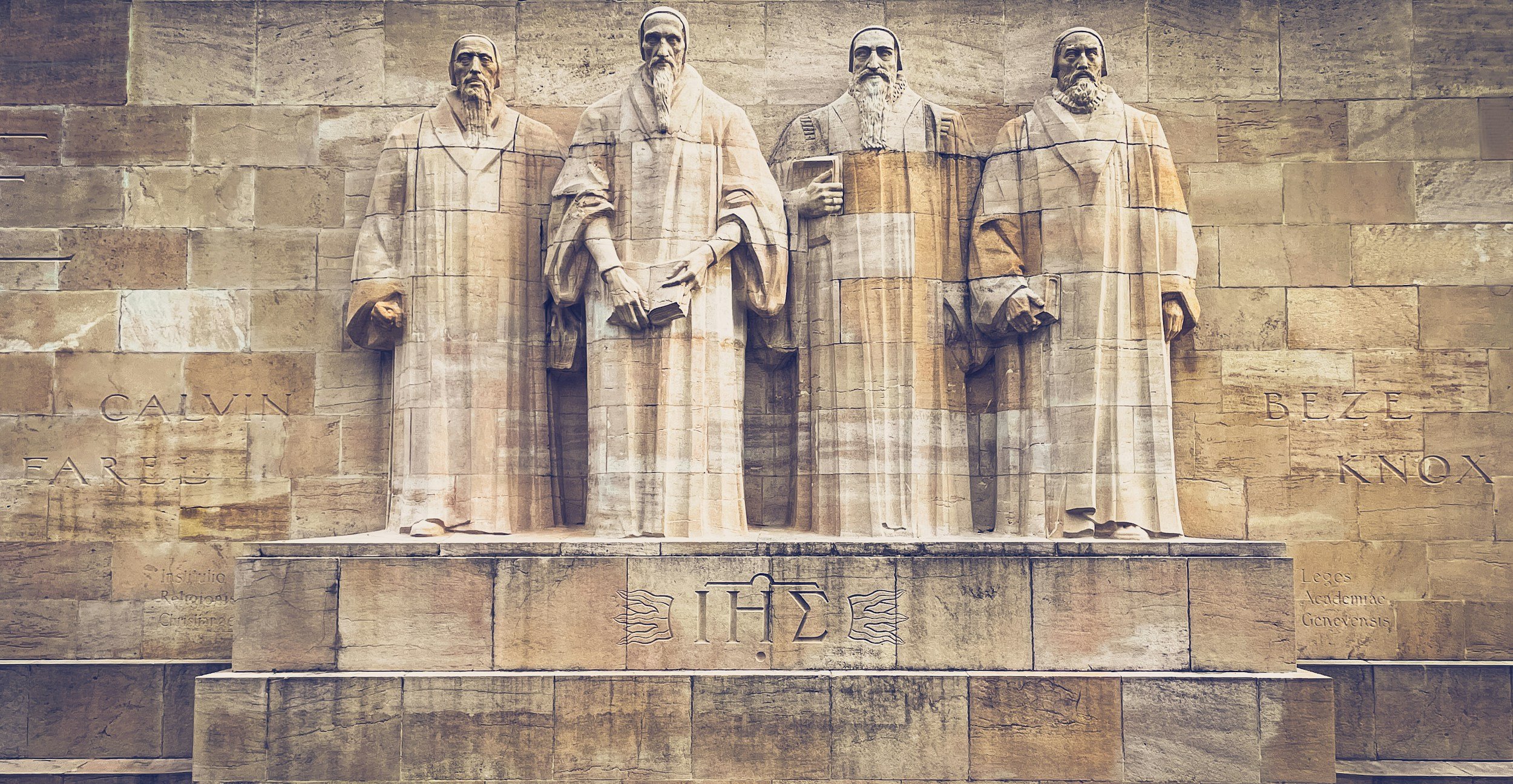 Statue of four historical figures on a stone wall, with inscriptions in French and Greek. The figures are dressed in robes and hold books or scrolls.