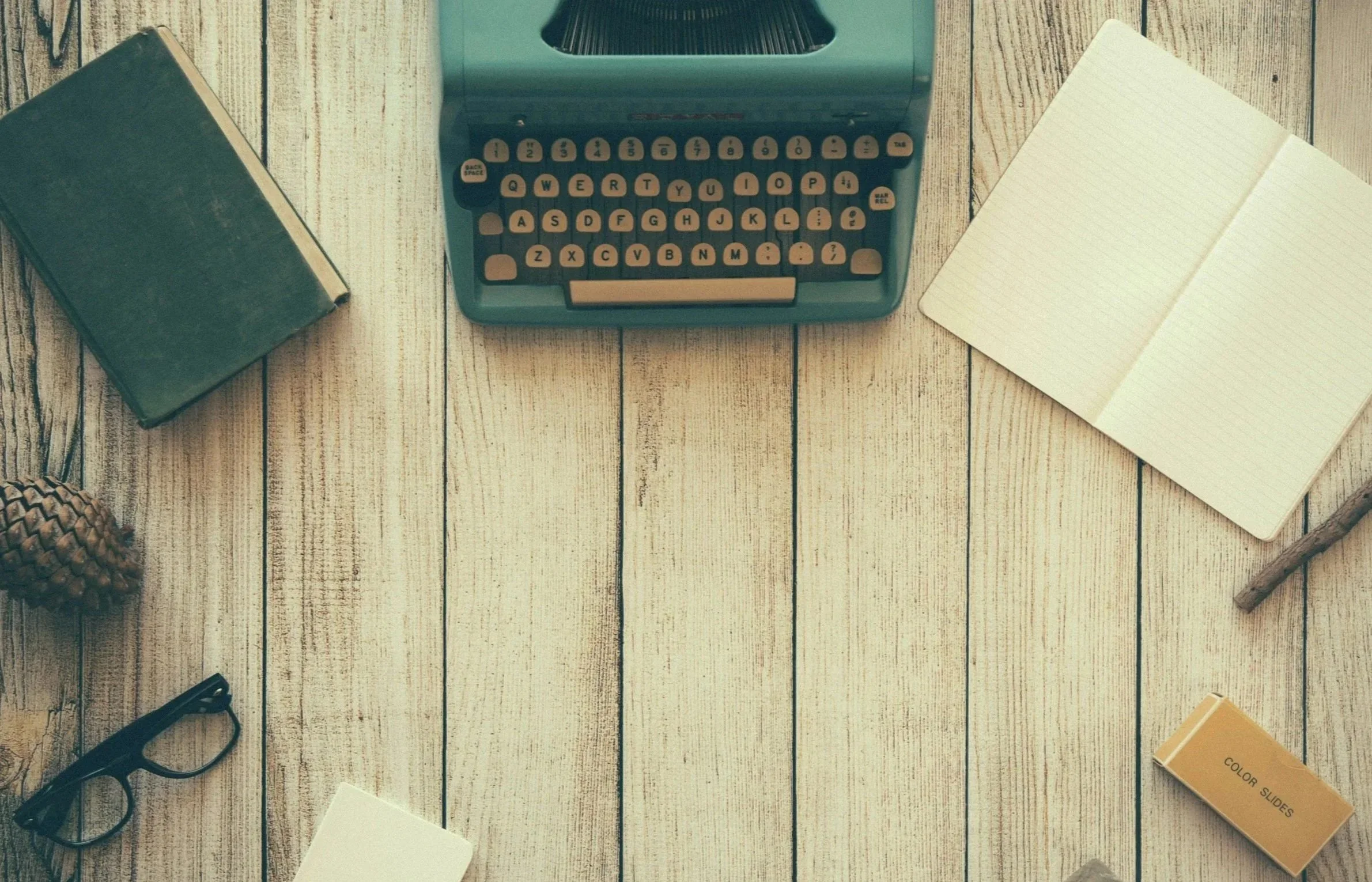 A vintage typewriter, an open notebook, a closed book, a pinecone, a pair of glasses, a sticky note, a small box, and a pencil on a wooden surface.