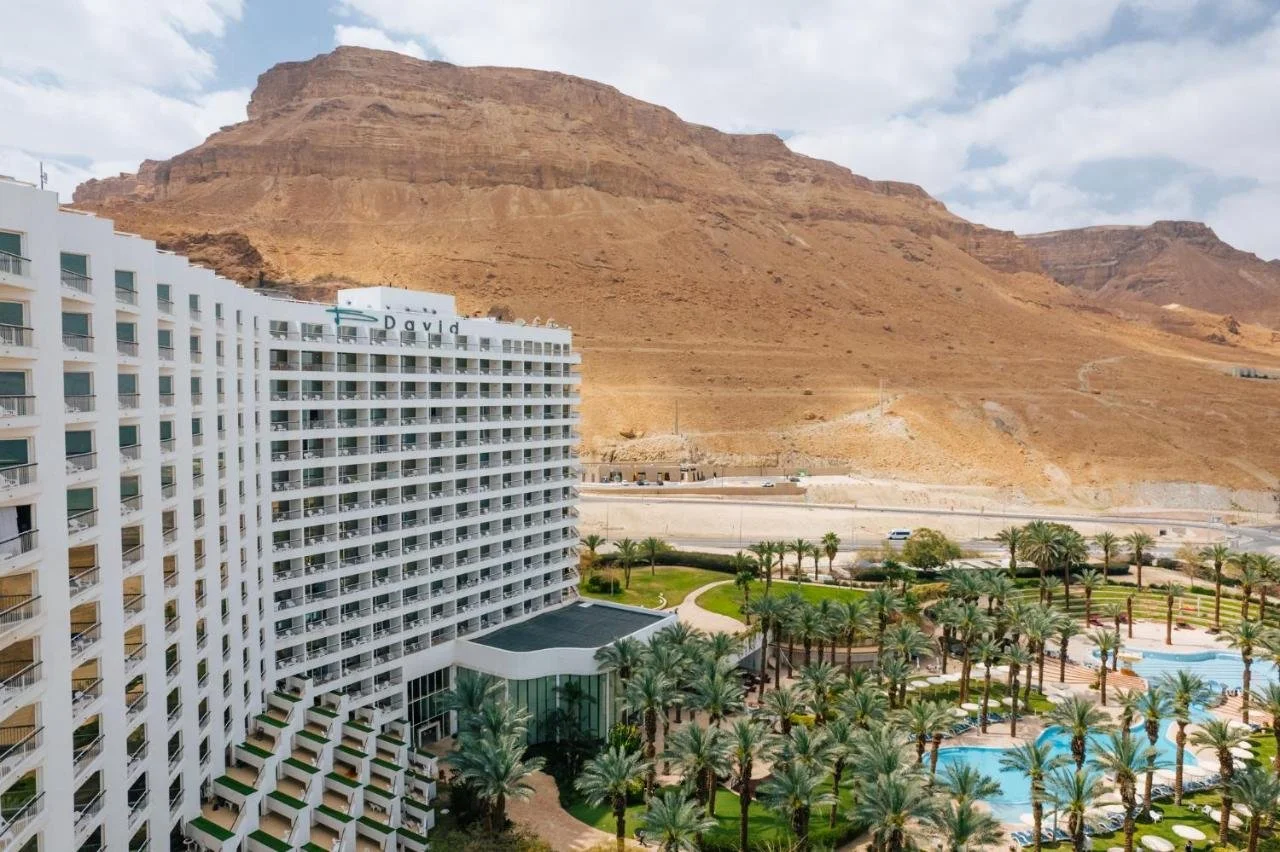 A tall white hotel building with many balconies surrounded by palm trees, with a mountain in the background and a pool area in the foreground.