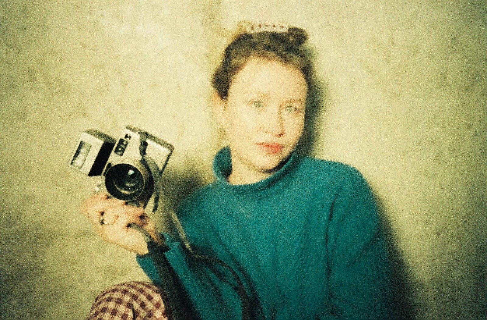 A woman with short hair tied back with a bow, wearing a teal sweater, holding a vintage camera, sitting against a beige textured wall.