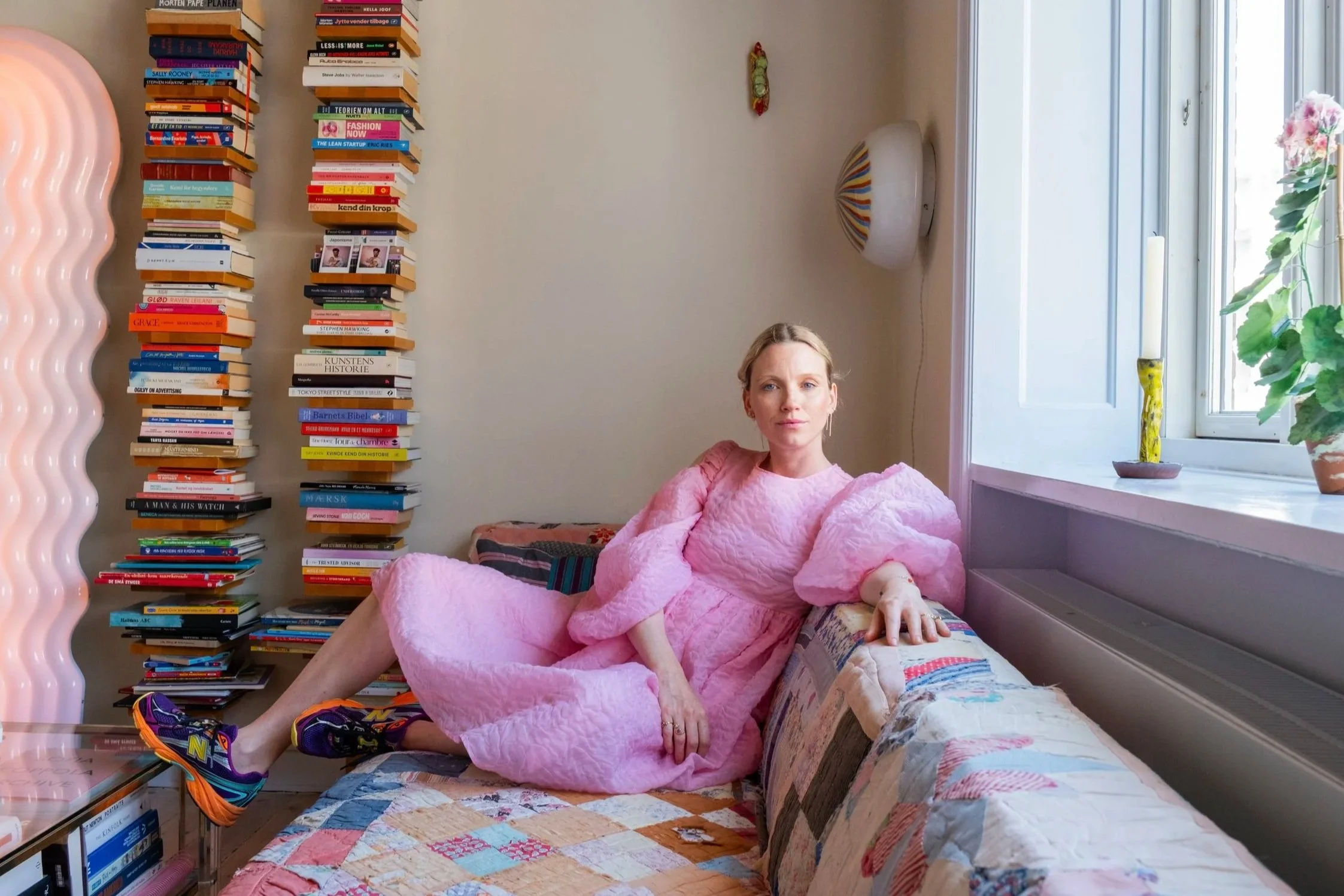 A woman in a pink dress and colorful sneakers lounging on a couch next to a tall stack of books in a living room with a large window and potted plant.