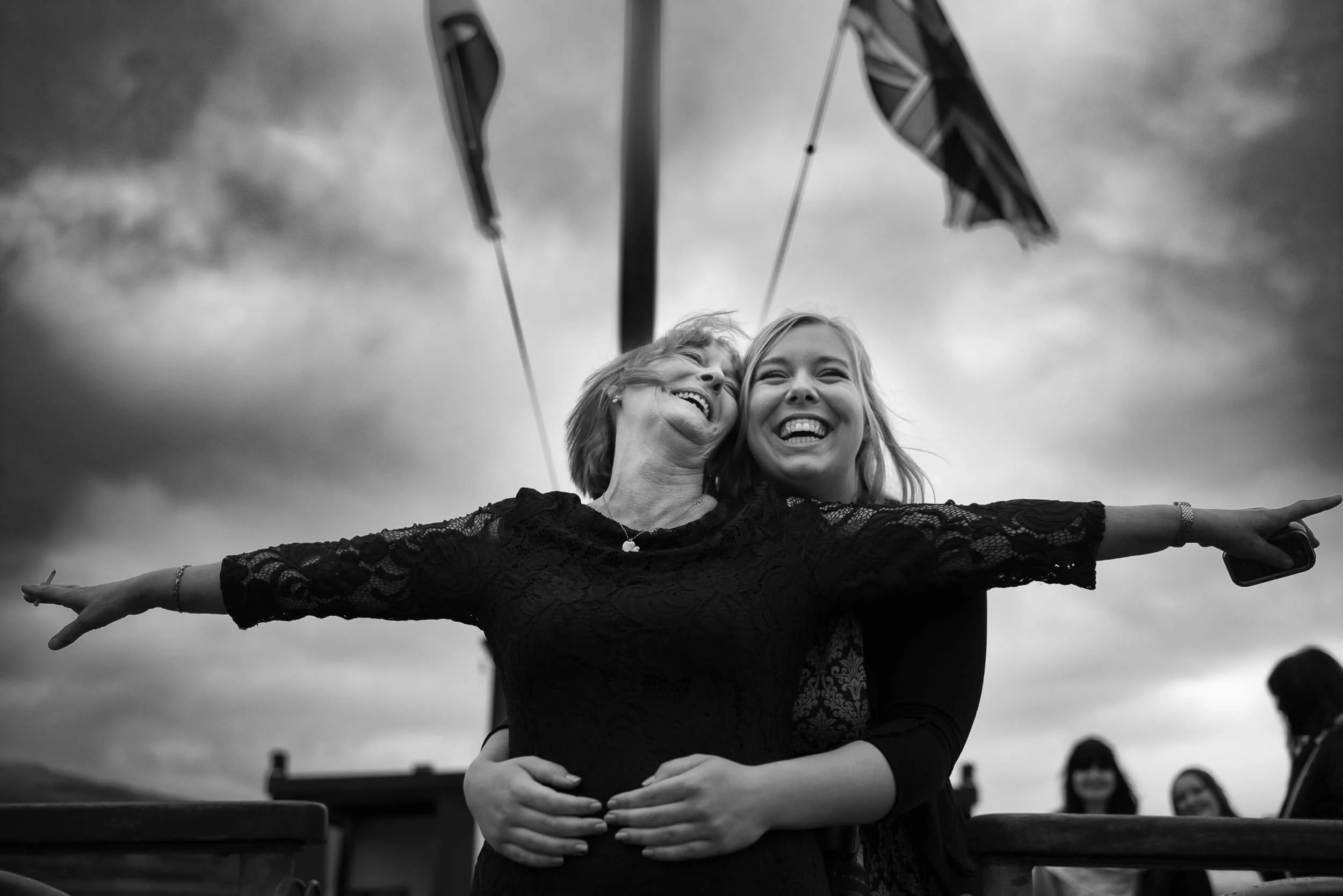 Two women are smiling and hugging each other outdoors, one woman extending her arms outward. Behind them are flags and a cloudy sky, with a few people blurred in the background.