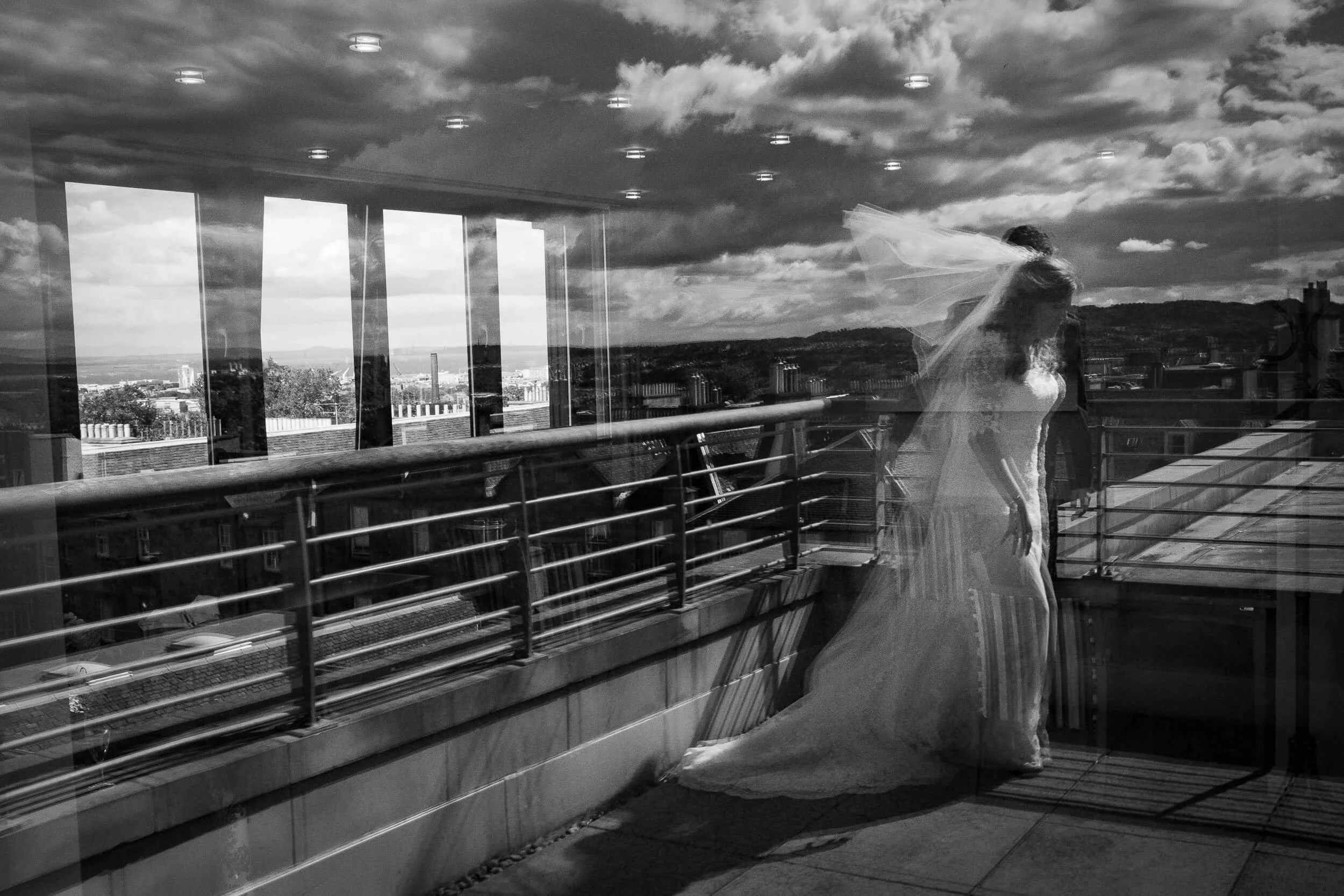 Reflection of a woman in a wedding dress on a glass window with cityscape and cloudy sky visible behind her.