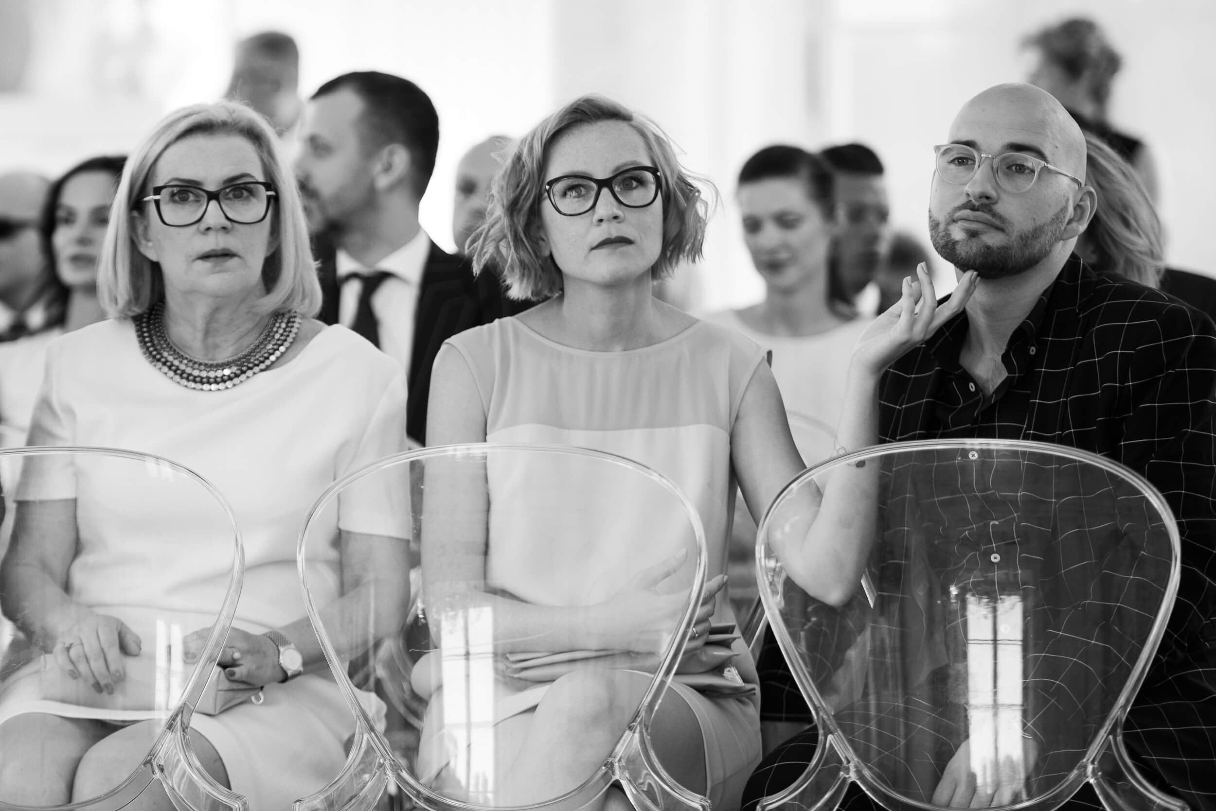 Black and white photo of a group of people sitting in an audience, looking attentive. Three women in the front row, all wearing glasses, with serious expressions. Behind them are various other individuals also seated and focused.
