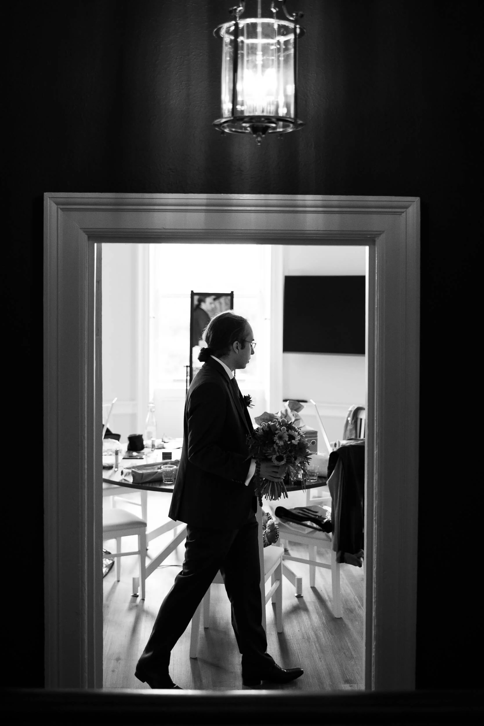 A man dressed in a suit holding a bouquet of flowers, seen through a doorway, with a bright, well-lit room behind him.