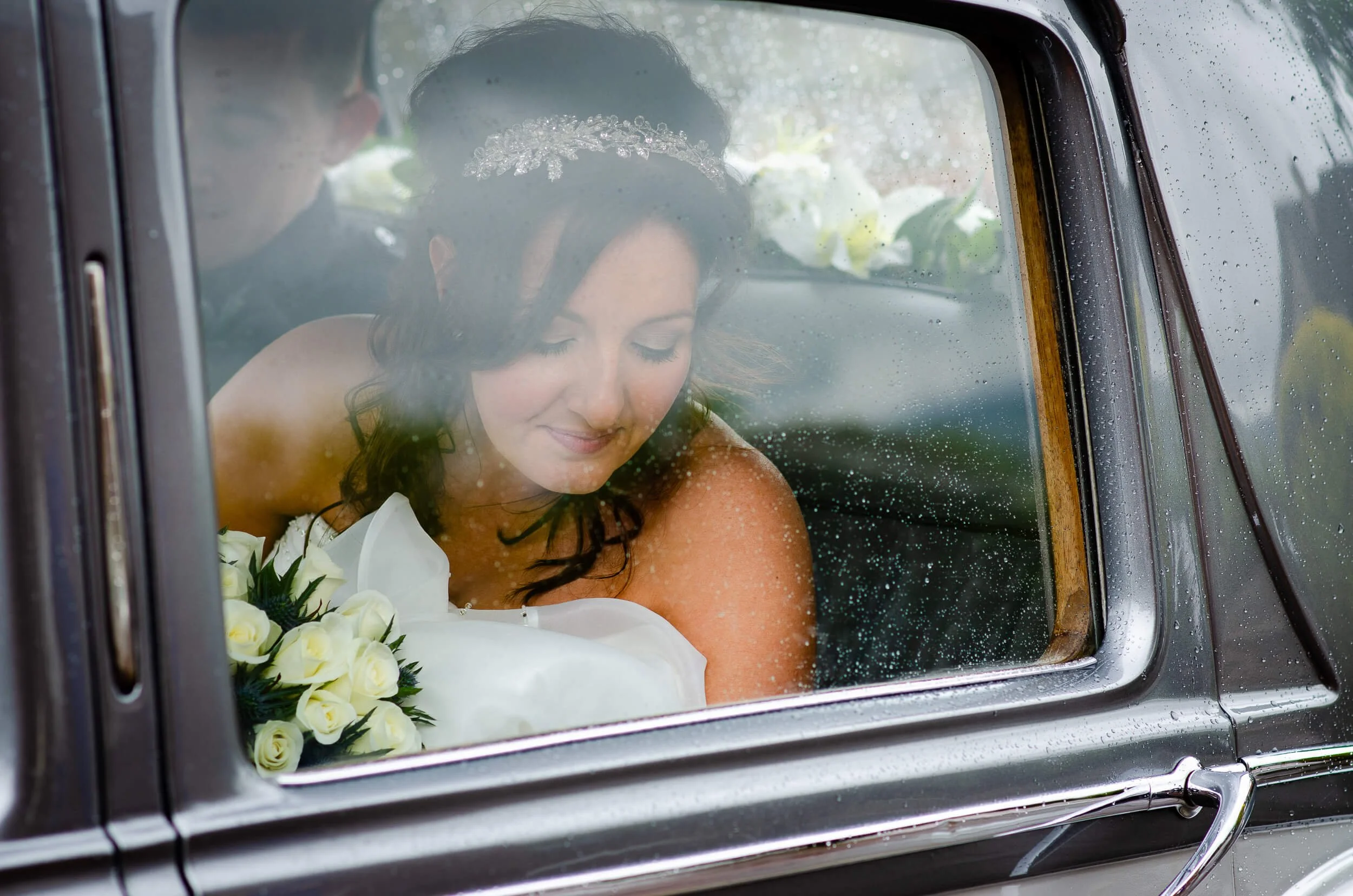 Bride sitting inside a vintage car, holding a bouquet of white roses, with a veil and floral headpiece, seen through a rain-spattered window.