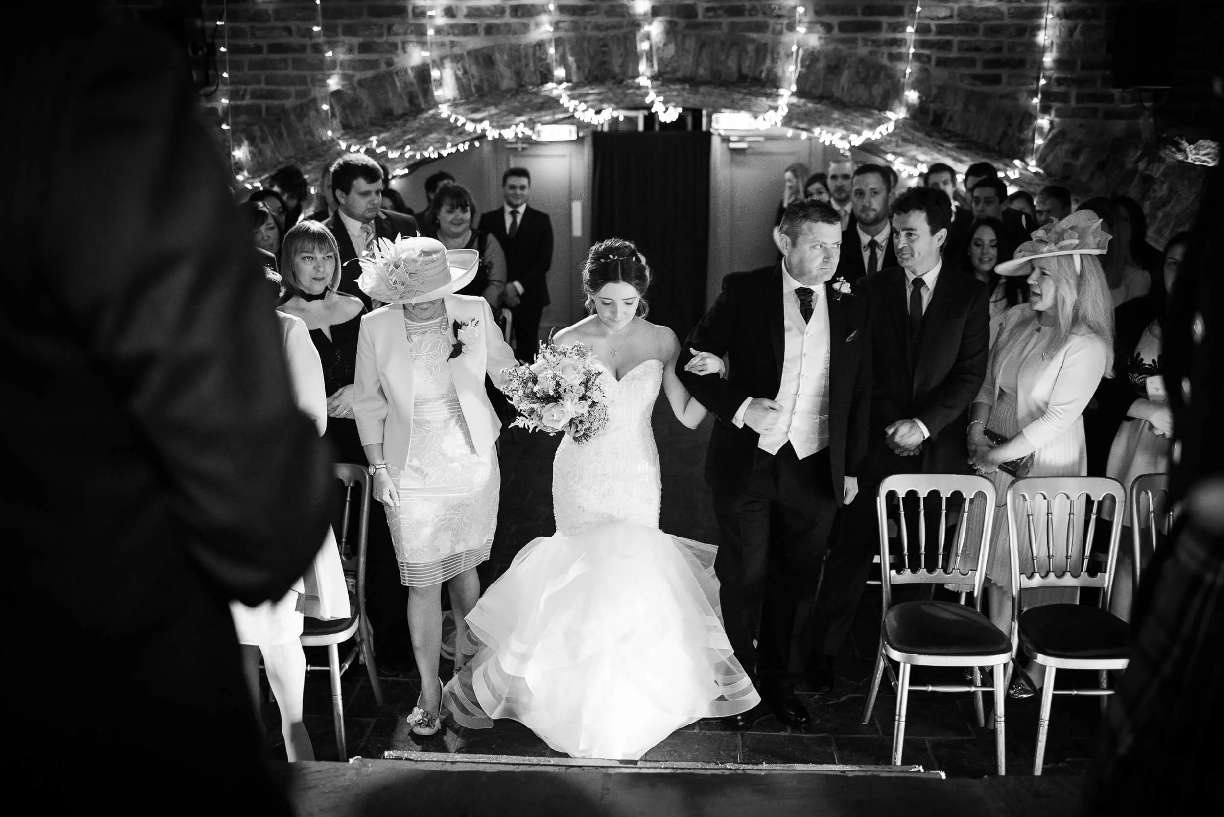 A black-and-white photo of a wedding ceremony with the bride in a strapless gown holding a bouquet, walking down the aisle with her arm linked to a groom dressed in a tuxedo. Family and friends are standing on either side, with some wearing hats, und