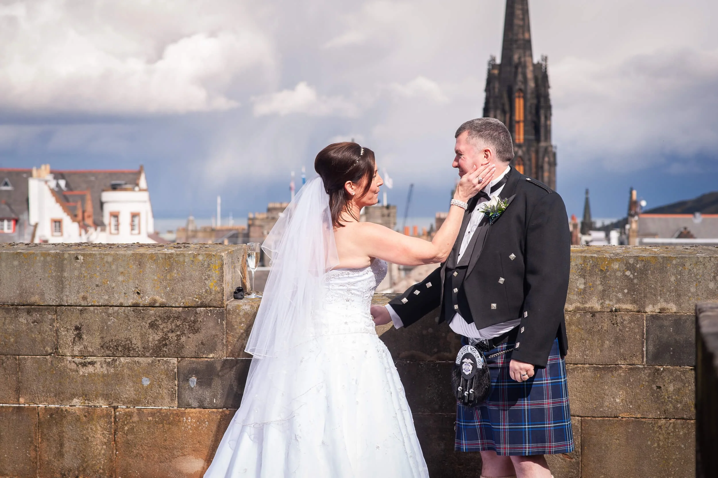 Bride touching groom's face on Edinburgh Castle ramparts with Edinburgh skyline behind — documentary wedding photography Scotland