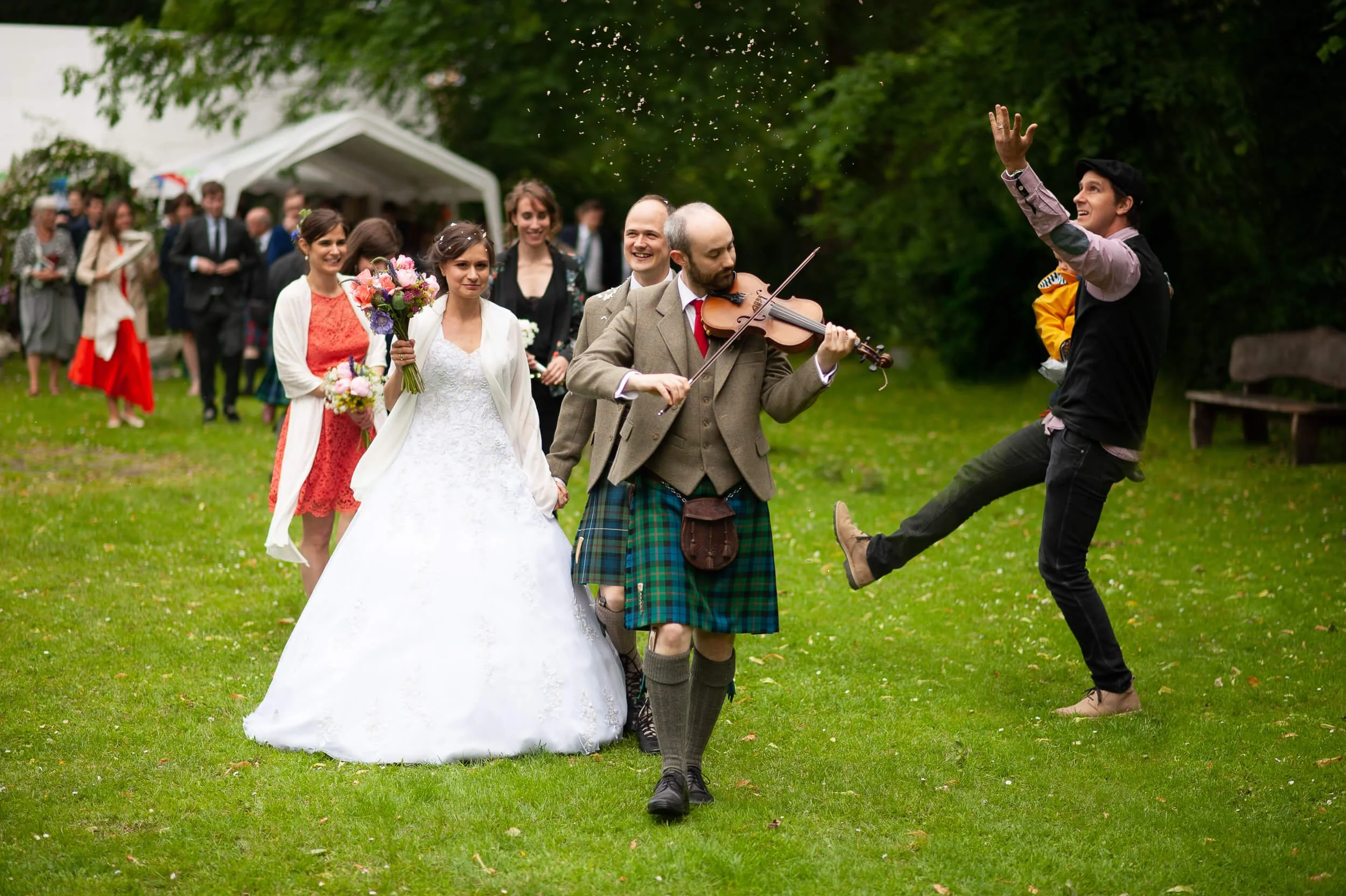 A wedding procession with a bride in a white wedding dress and veil, flowers, and several guests, with a musician playing a violin and a man dancing energetically in a kilt and jacket on a grassy outdoor area surrounded by trees.