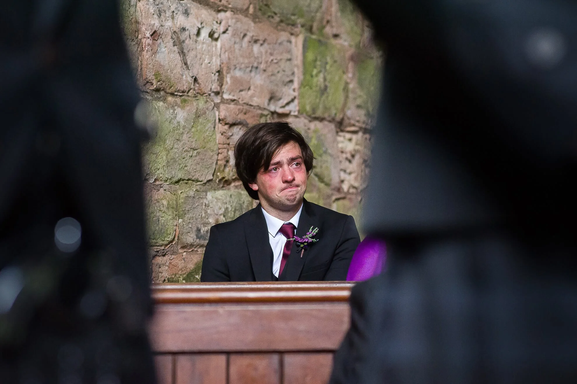 A young man with dark hair, wearing a suit and tie, sitting against a brick wall. He has a somber expression with tears in his eyes.