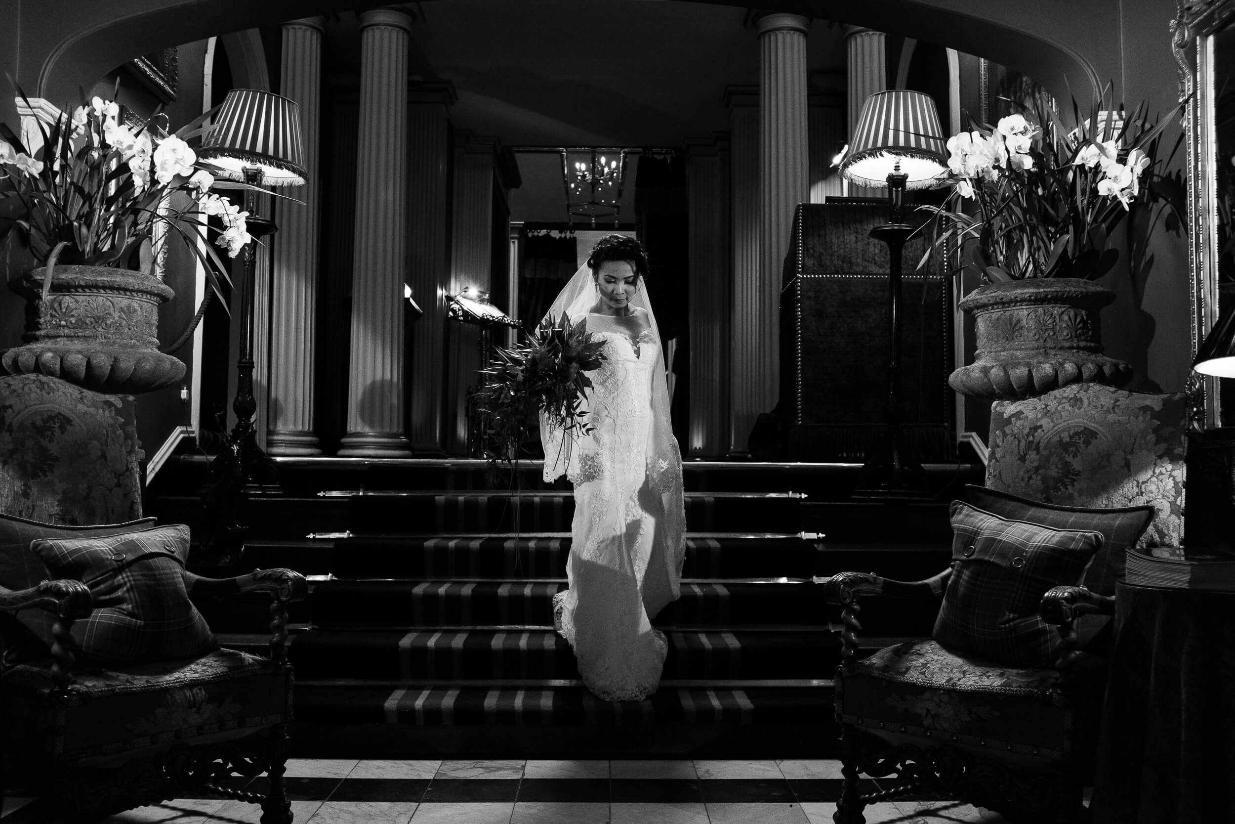 A bride in a wedding dress and veil, holding a bouquet, walking down a staircase in an ornate, elegantly decorated interior.