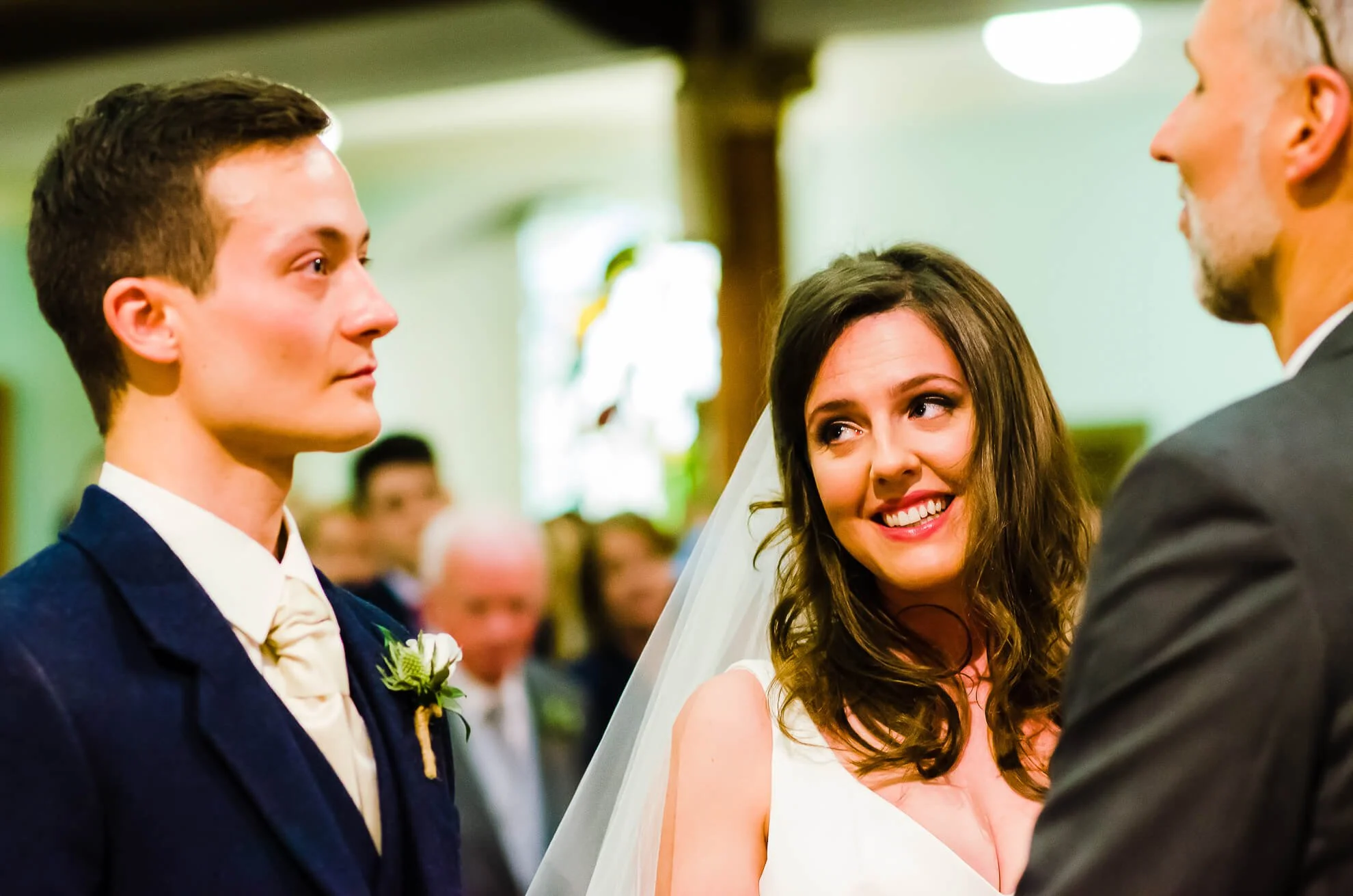 Bride and groom looking at each other during wedding ceremony in church, with a smiling officiant in the background.