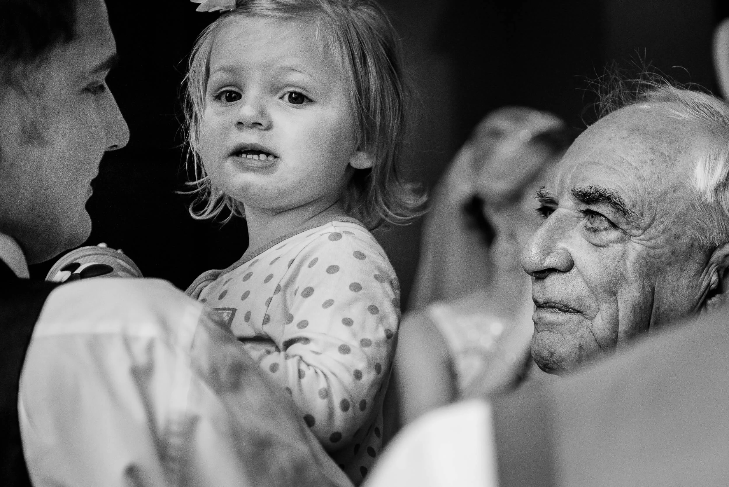A young girl with a flower in her hair, looking at an elderly man with a solemn expression, with other people in the background.