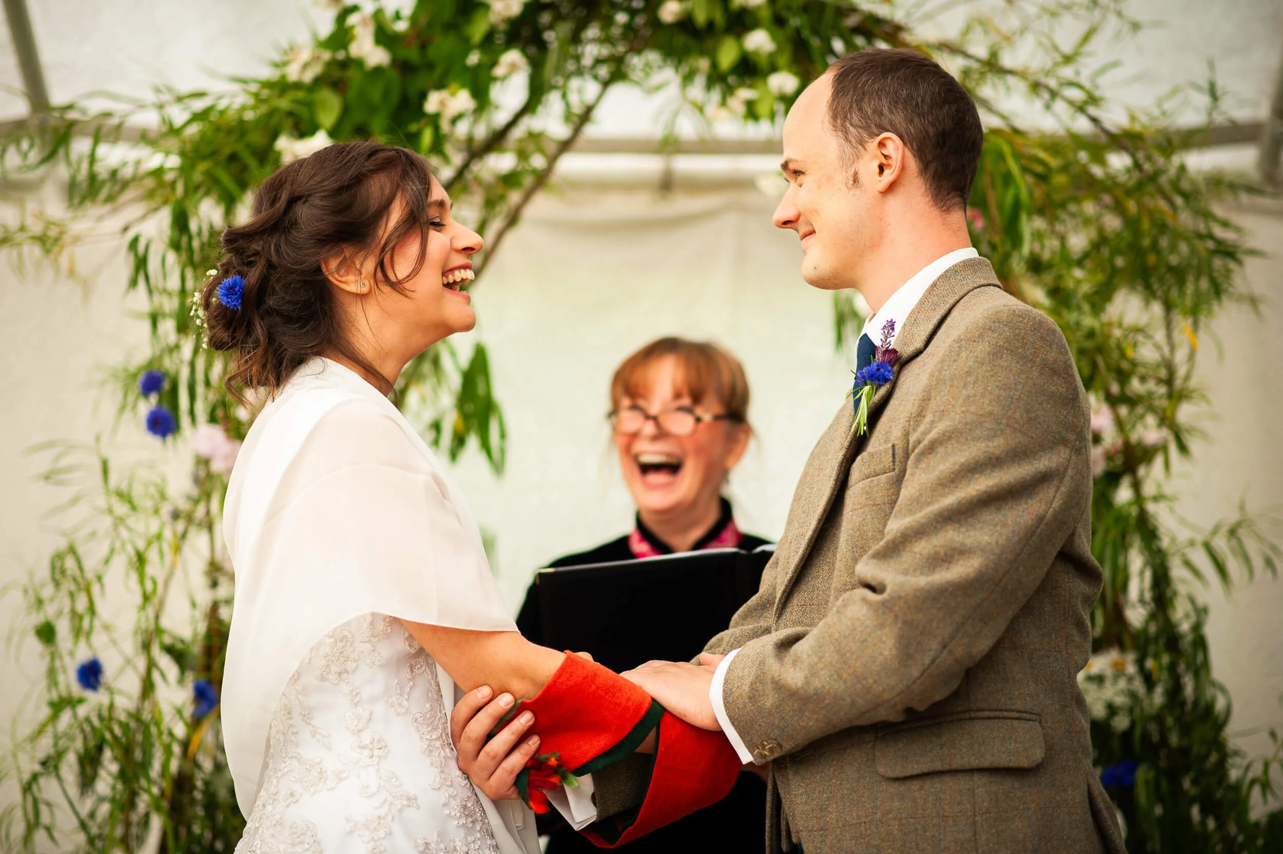 A couple is getting married in a wedding ceremony, holding hands and smiling at each other. The bride has dark hair with blue flowers and is wearing a white dress. The groom has short brown hair, wearing a brown jacket and a purple boutonniere. An of