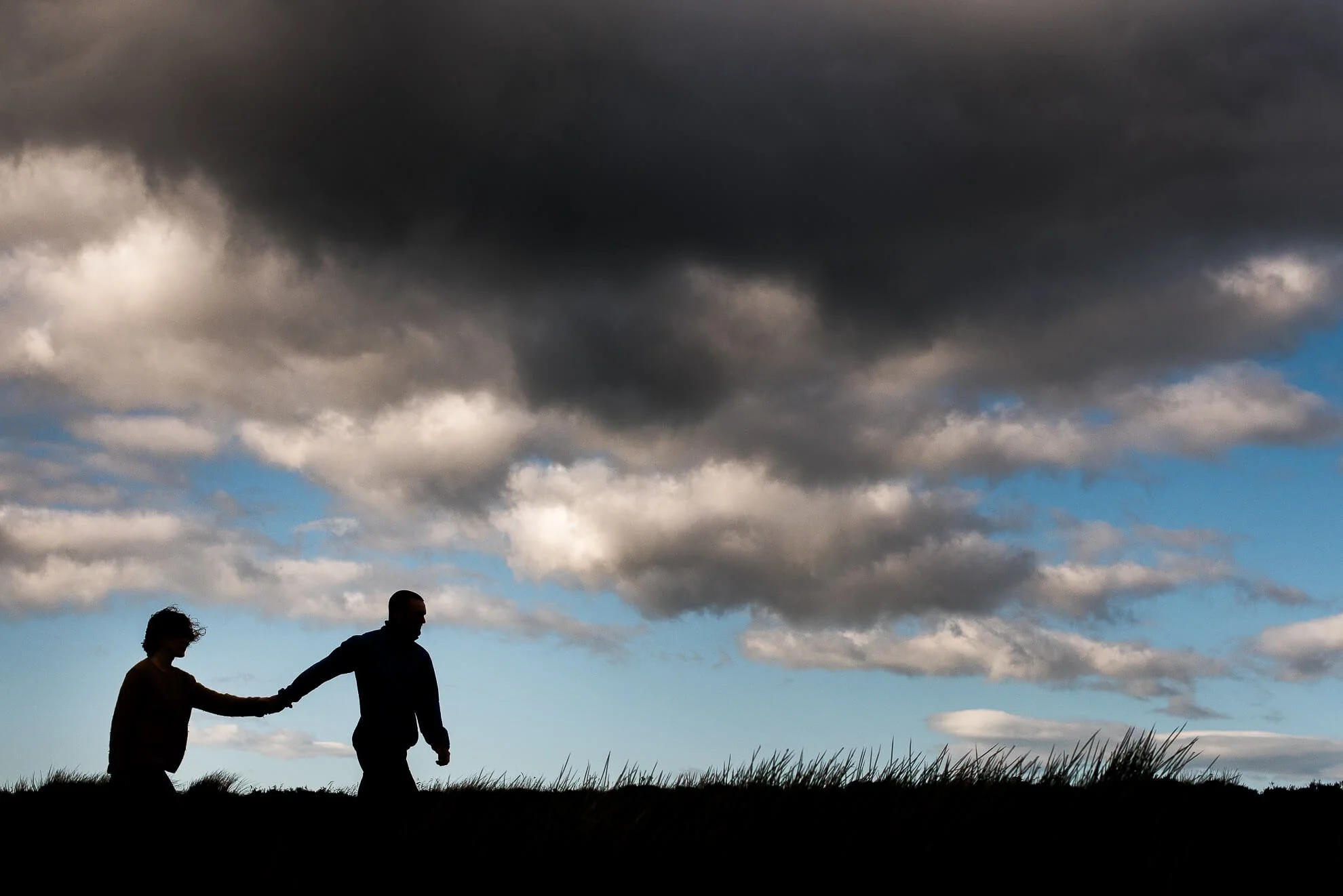 Silhouettes of two people holding hands walking outdoors under a dark, cloudy sky with patches of blue.