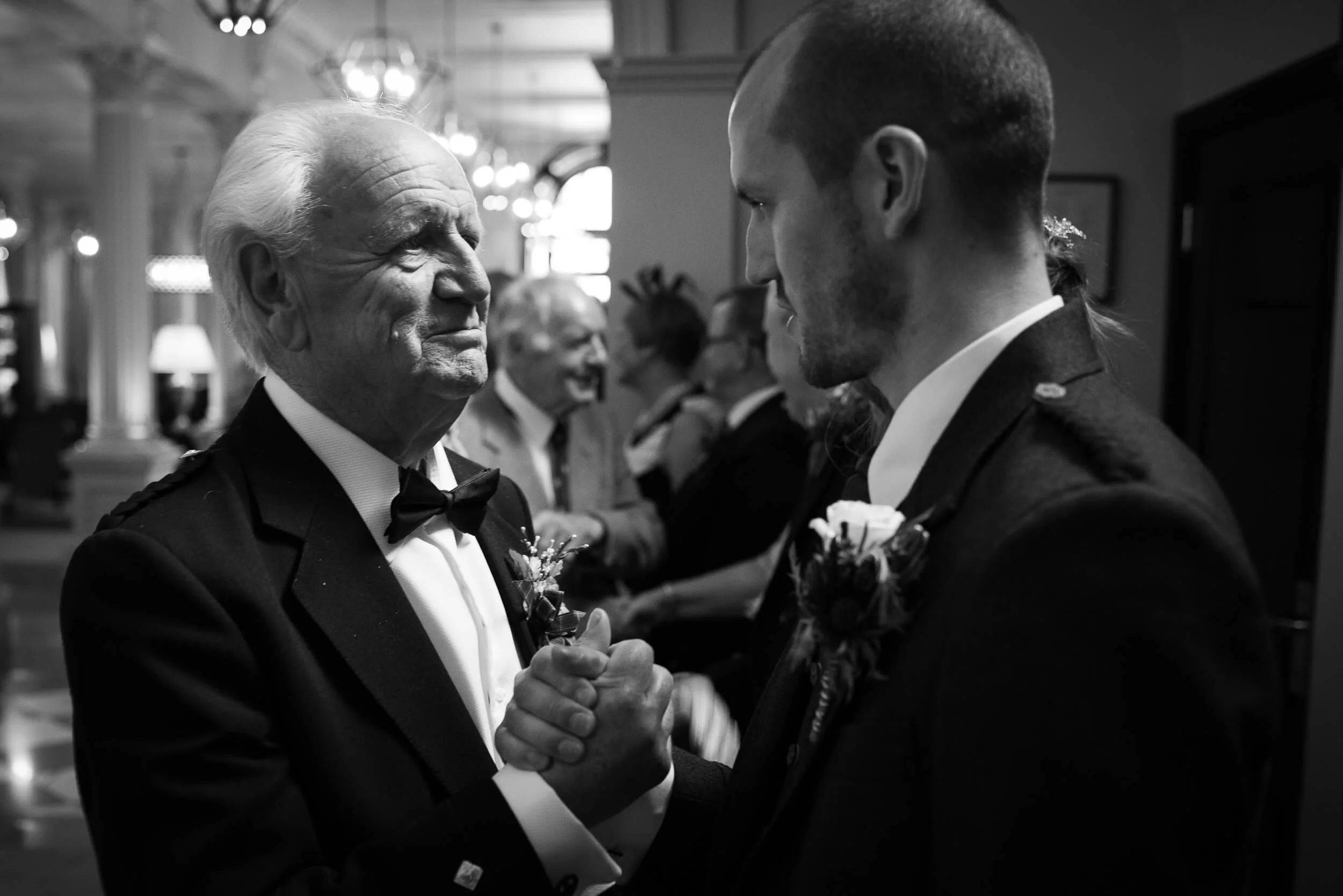An elderly man and a young man in formal tuxedos holding hands and looking at each other, likely at a wedding reception.