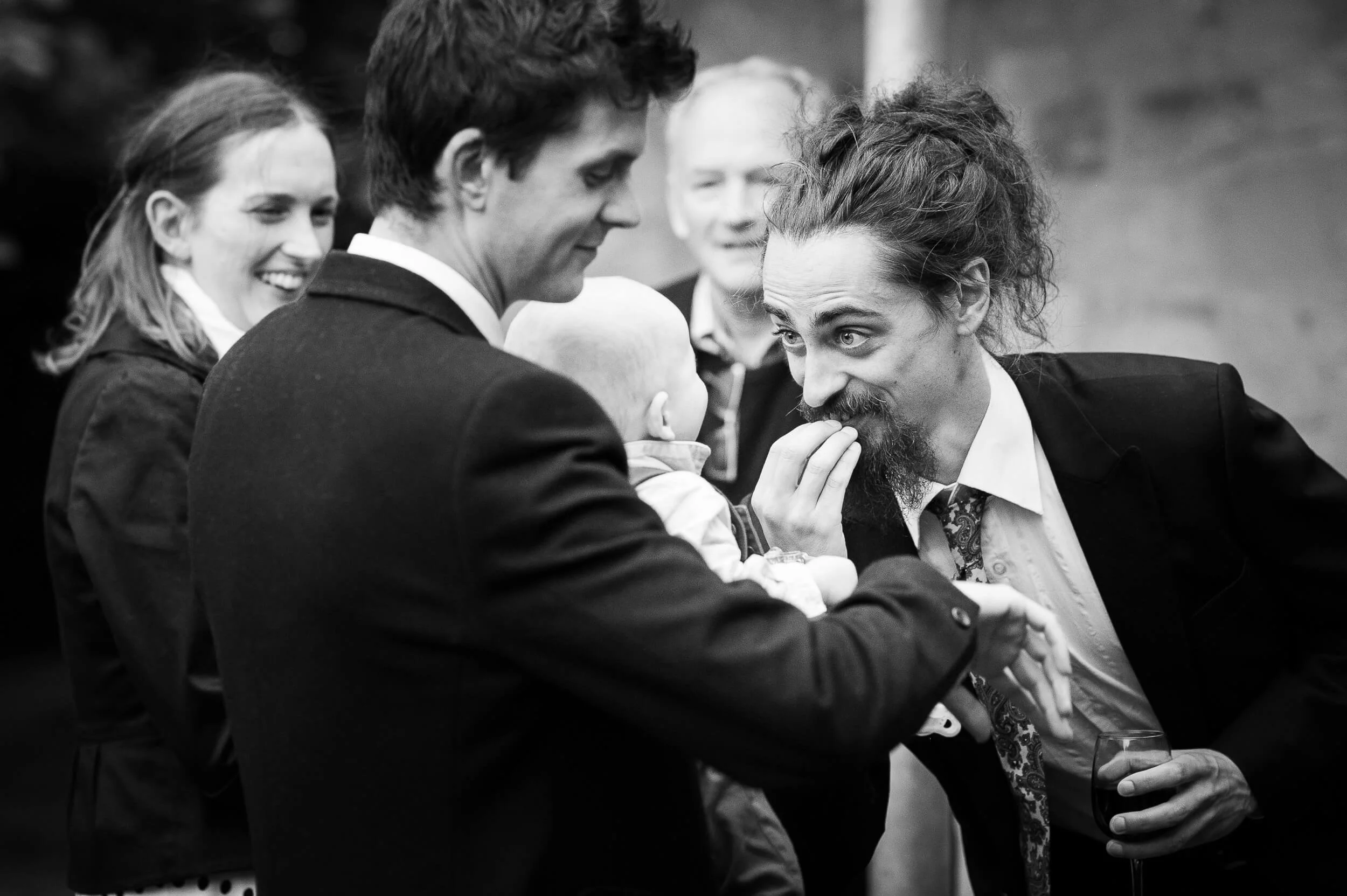 A group of people, including a man with long curly hair and a beard, engaging in conversation at a social gathering. One man is holding a baby and the others are smiling and talking, with one holding a glass of wine.