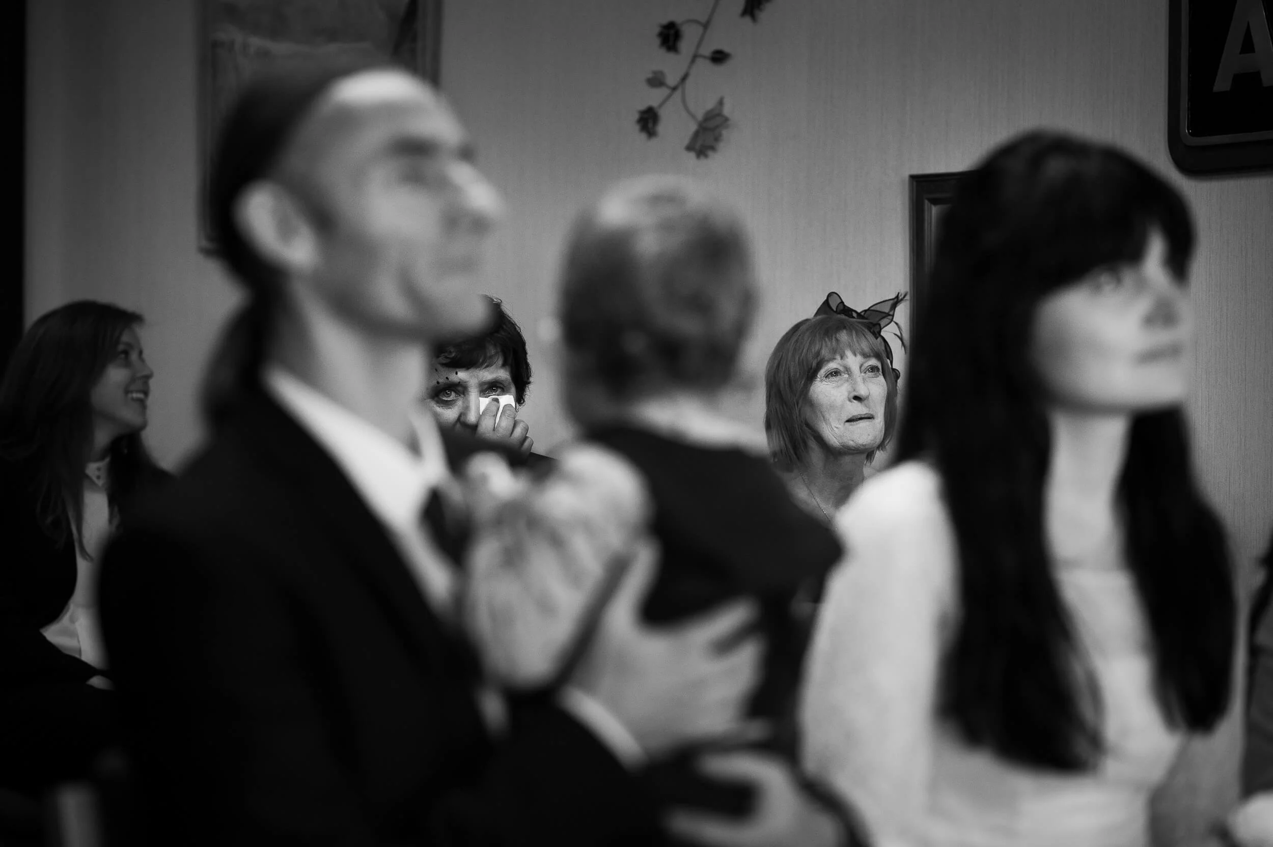 Black and white photo of people sitting in a room, with some individuals in the foreground out of focus and a woman in the background wiping her eyes or face.
