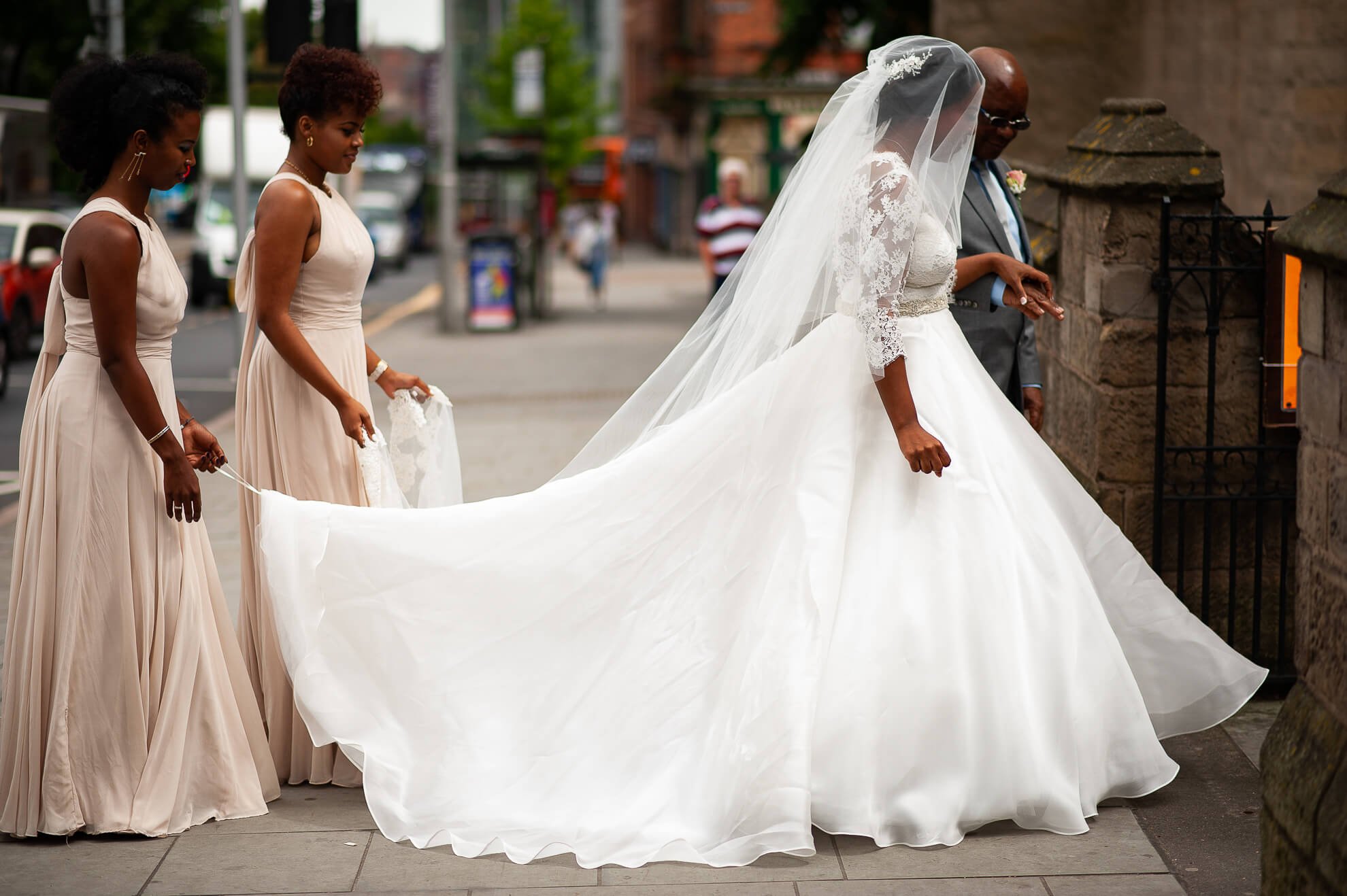 A bride in a white wedding gown and lace jacket walks with her bridesmaids in beige dresses on a city sidewalk. The bride wears a veil and holds her gown as she walks, with a man in a gray suit behind her.