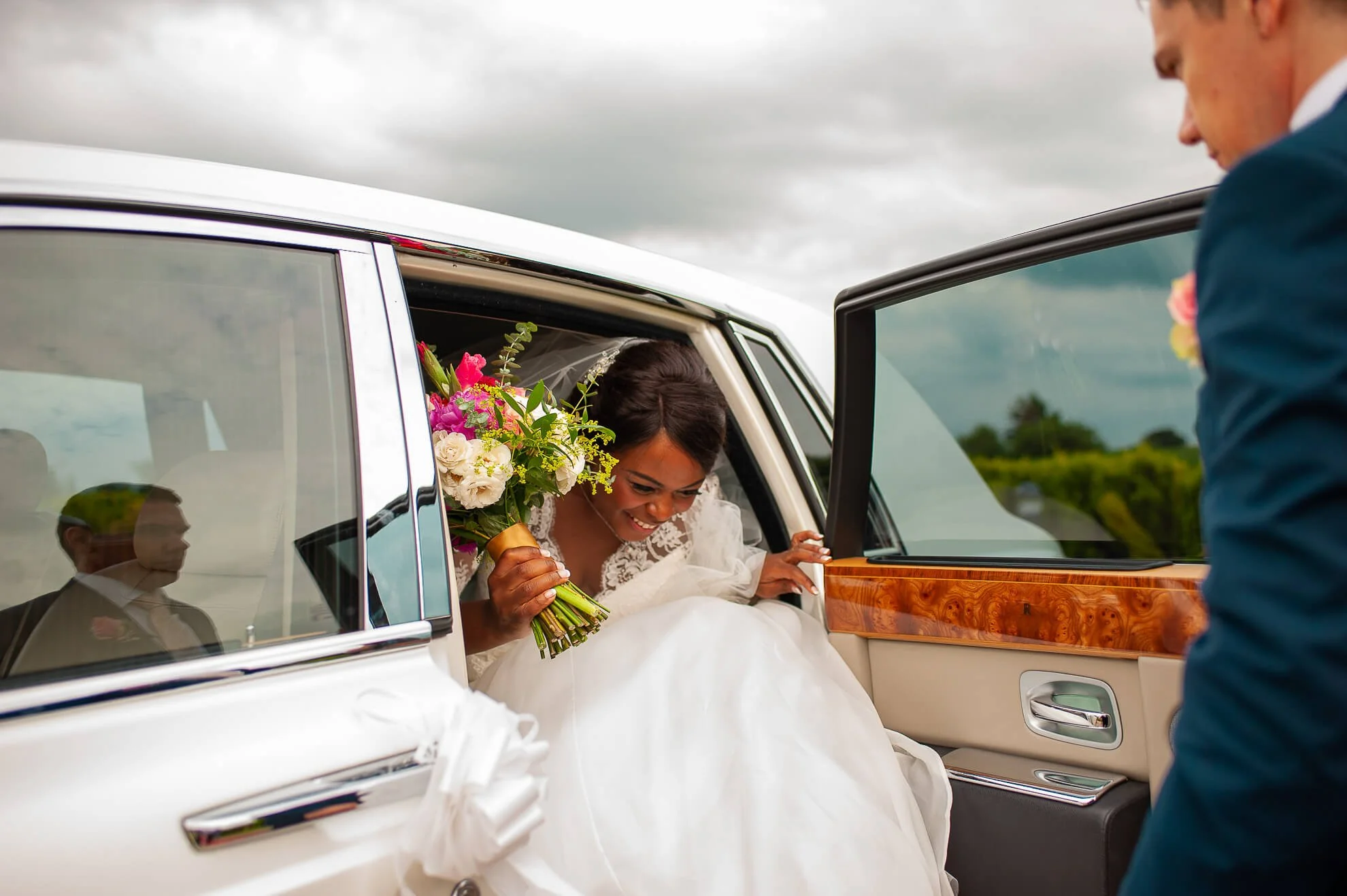 Bride with a bouquet of colorful flowers getting into a white luxury car, while a man in a blue suit stands nearby, under a cloudy sky.