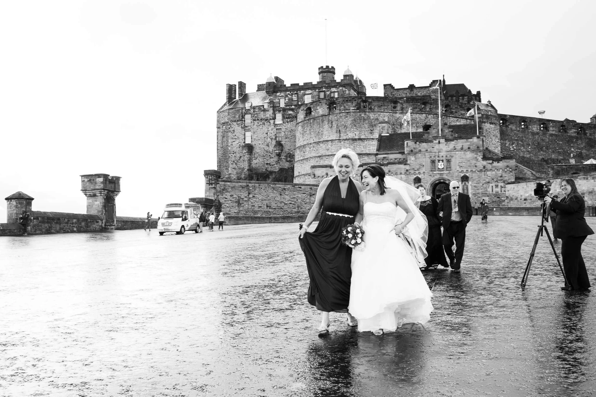 Bride and bridesmaid laughing on the wet esplanade of Edinburgh Castle — black and white documentary wedding photography Scotland